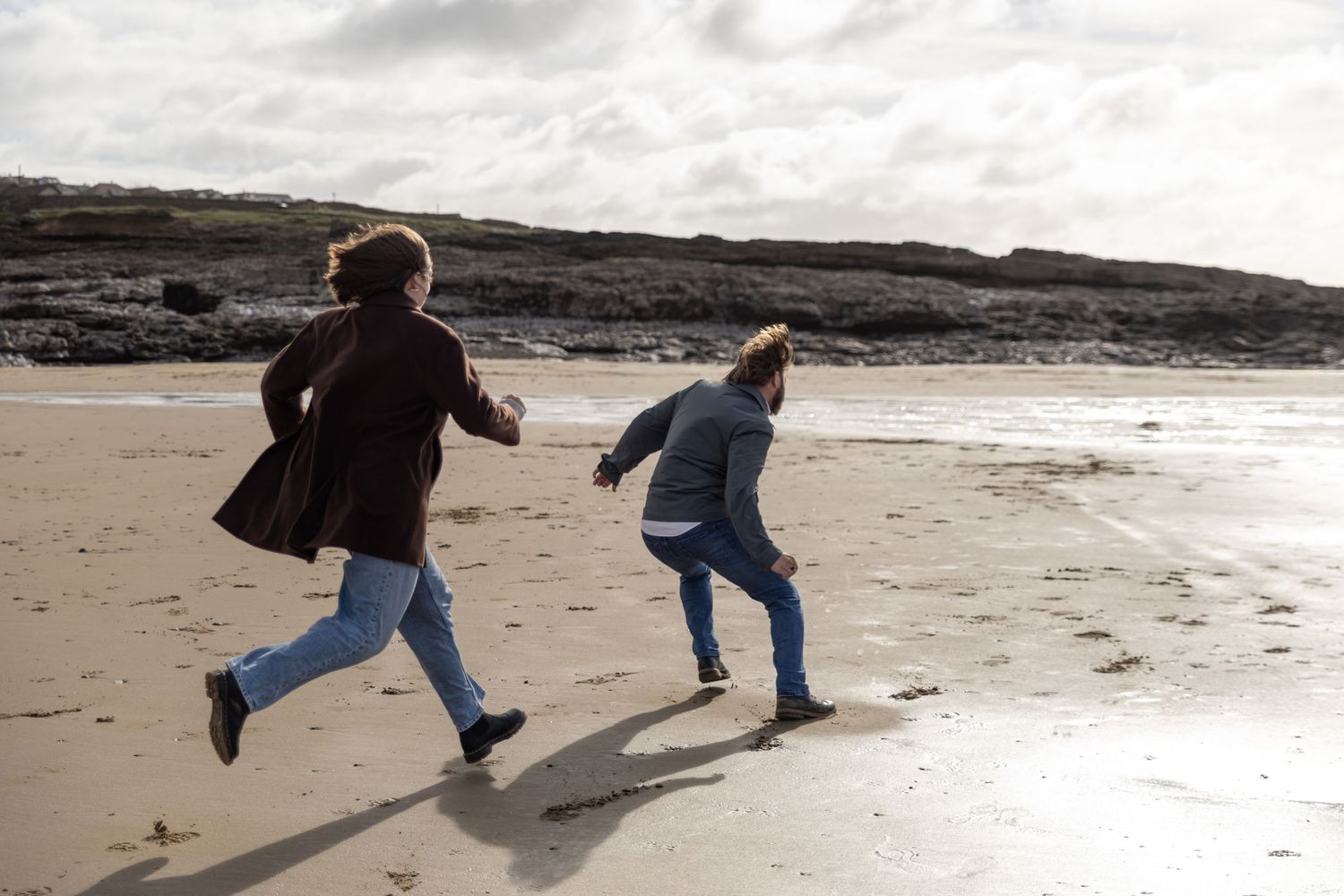 People run playfully across a sandy beach with cliffs visible in the background.