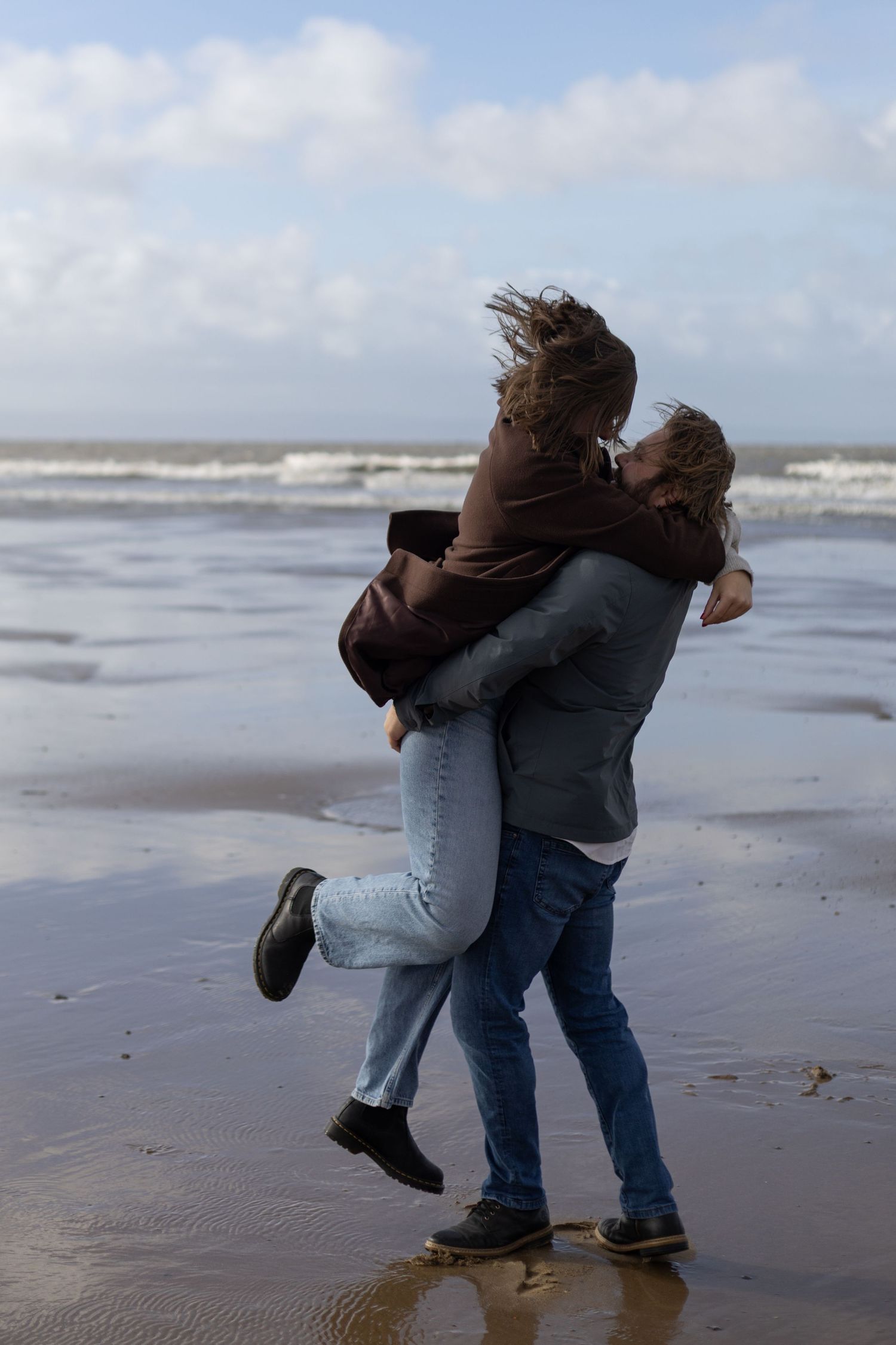 Two people playfully dance and embrace on a windswept beach.