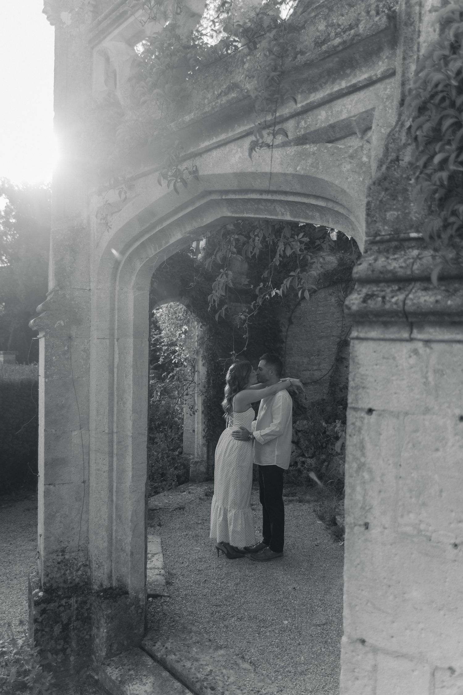 Black and white photo of a couple embracing under a stone archway with sunlight streaming through lush foliage.