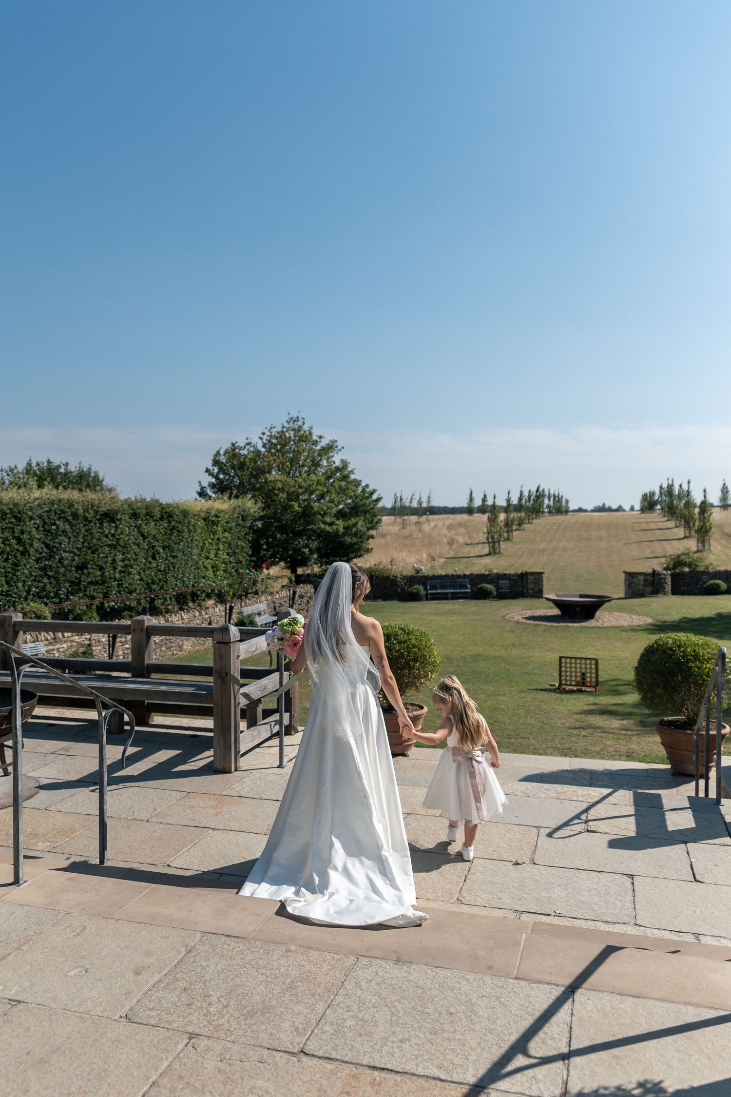 Bride in white gown and veil holds hands with flower girl on sunny patio, lush garden backdrop.