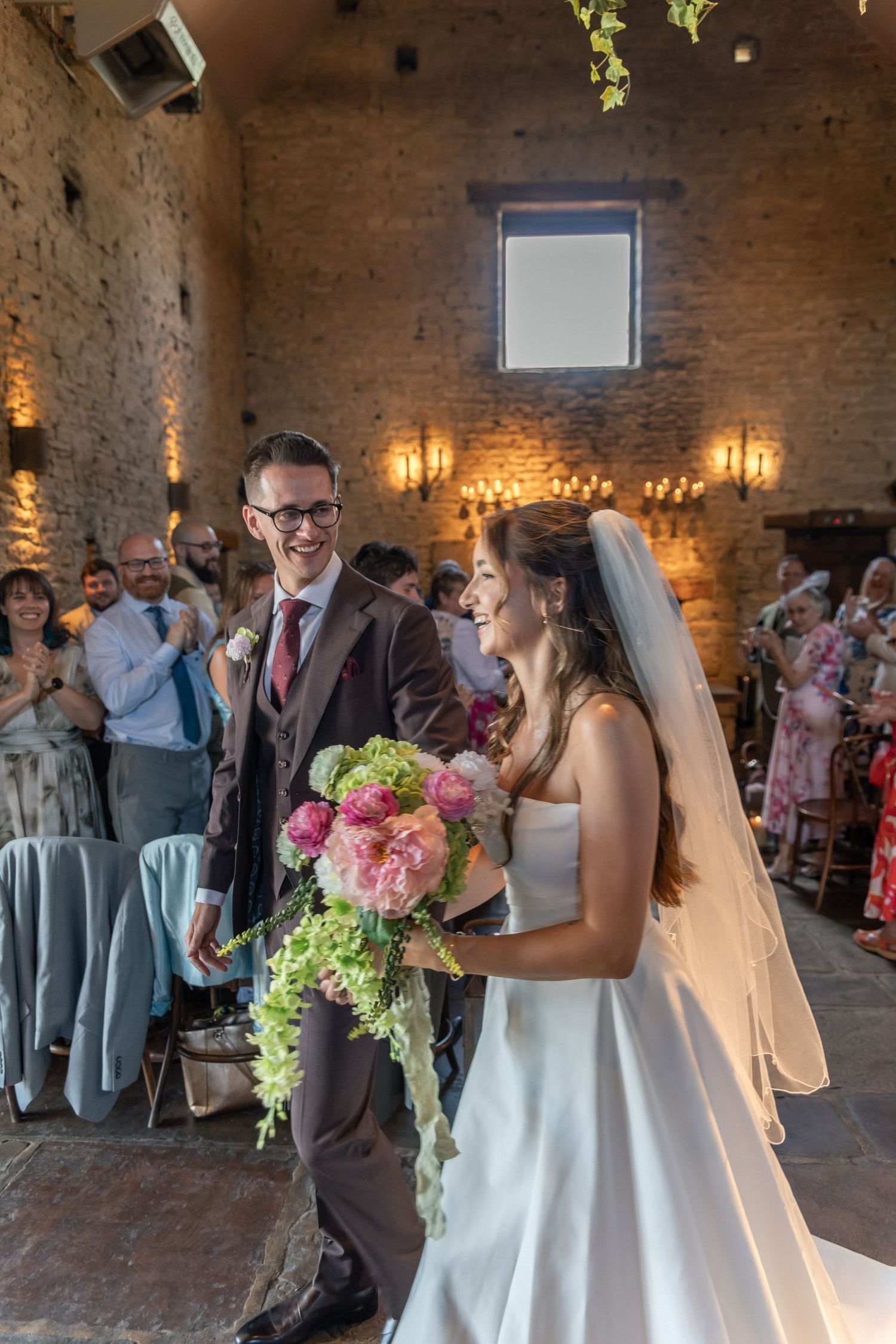 Joyful newlyweds walk down the aisle in a rustic stone venue, bride holding a cascading pink bouquet.