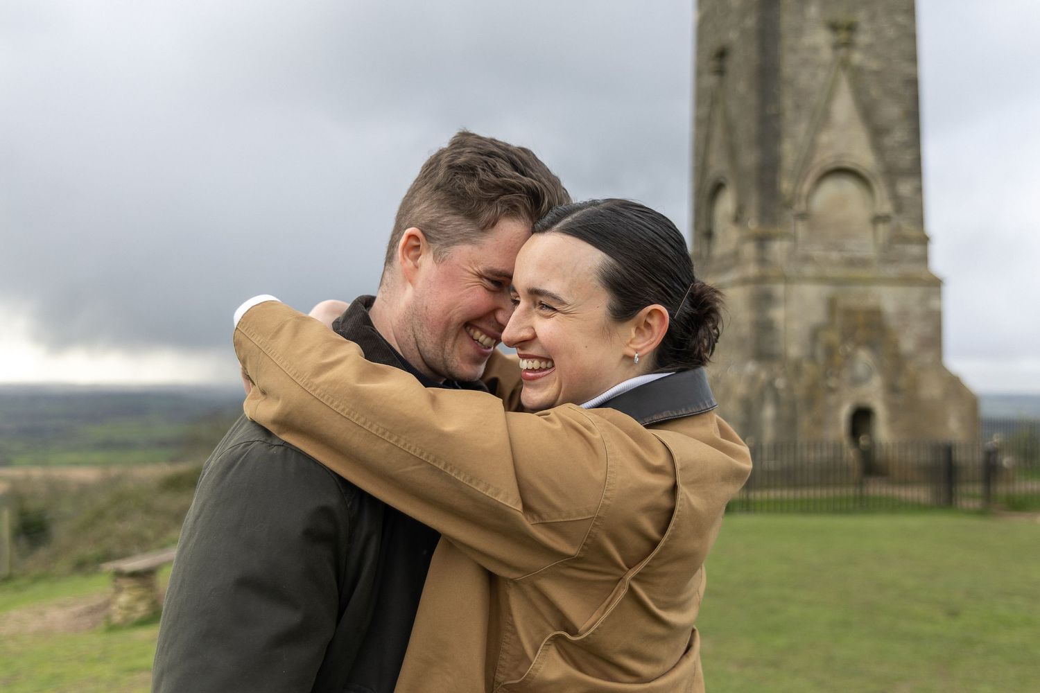 A couple embraces while laughing together near a stone tower monument in the countryside.