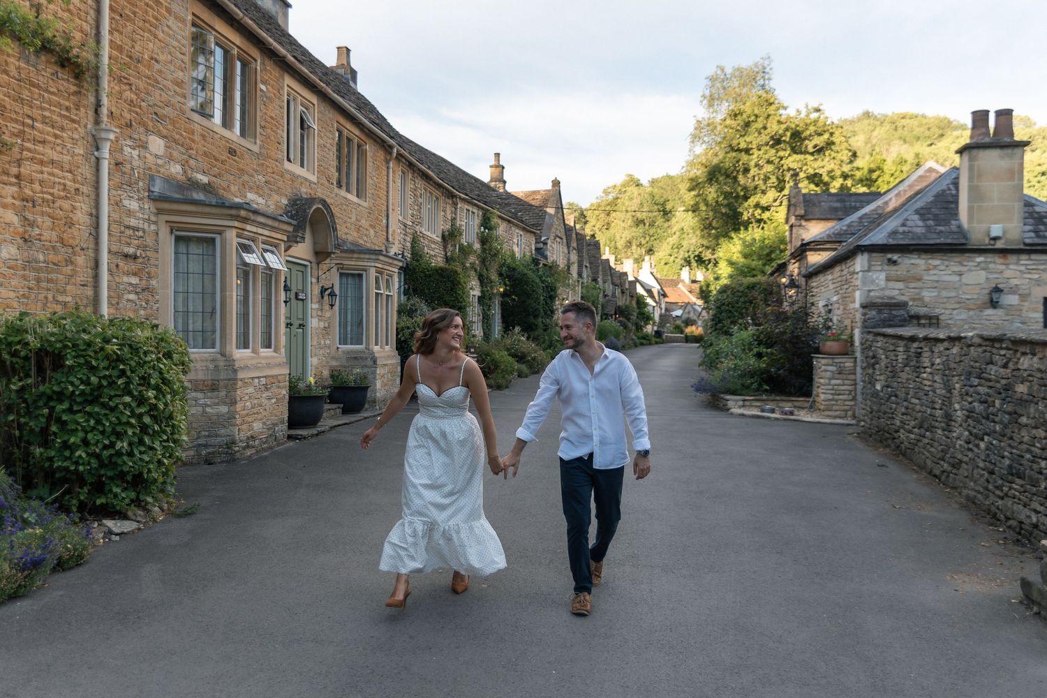 Couple in wedding attire holding hands and walking through a charming Cotswolds village street lined with stone cottages.