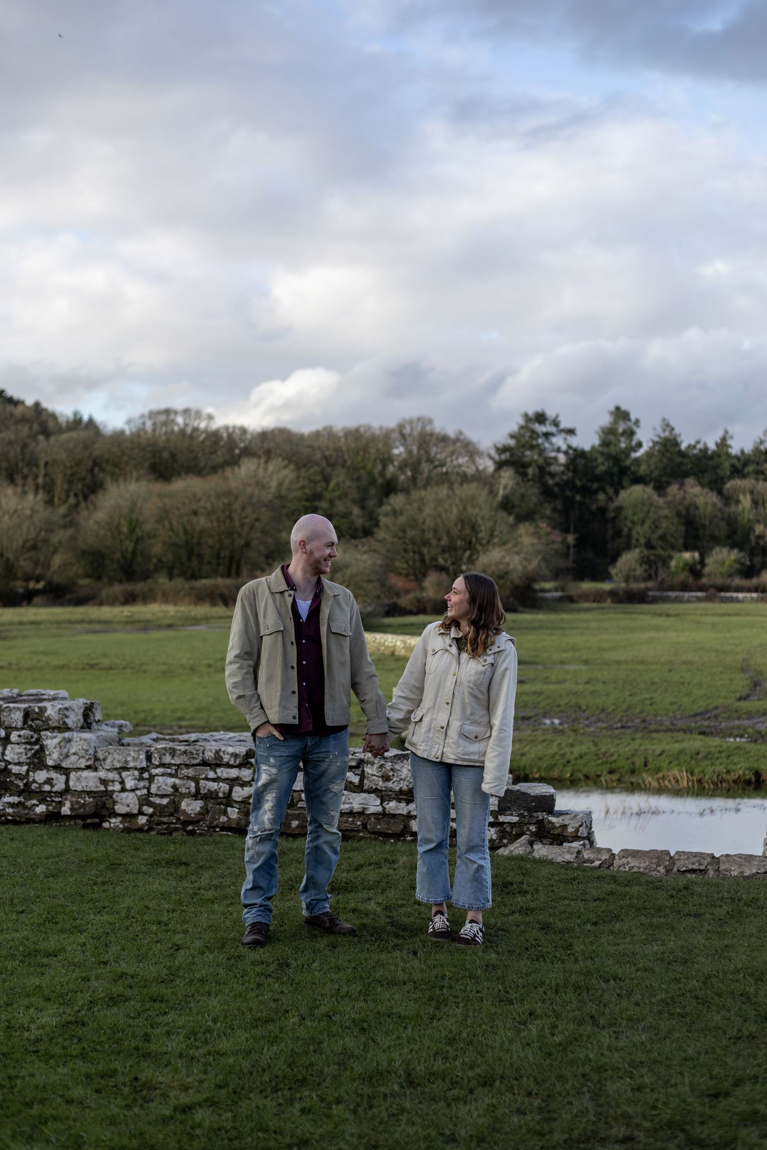 Two people stand together in a green field next to a stone wall on an overcast day.