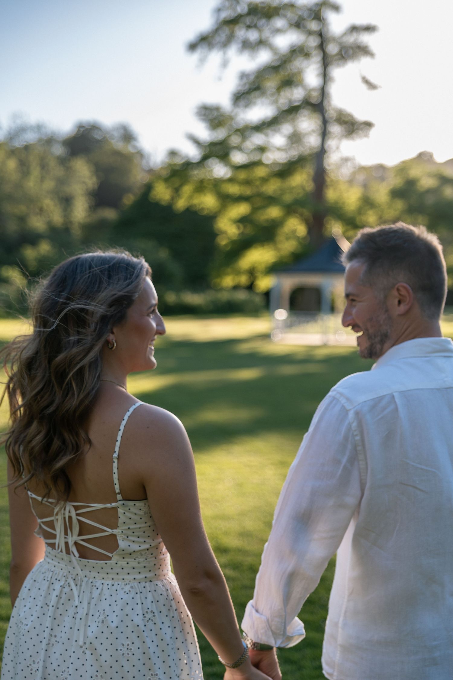 Couple holding hands in a sunny park, woman in white sundress looking back smiling, man in white shirt smiling at her.