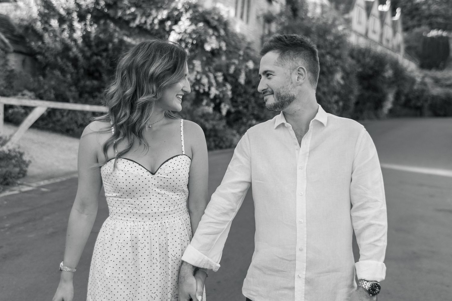Black and white photo of a smiling couple holding hands and walking together outdoors on a sunny day.