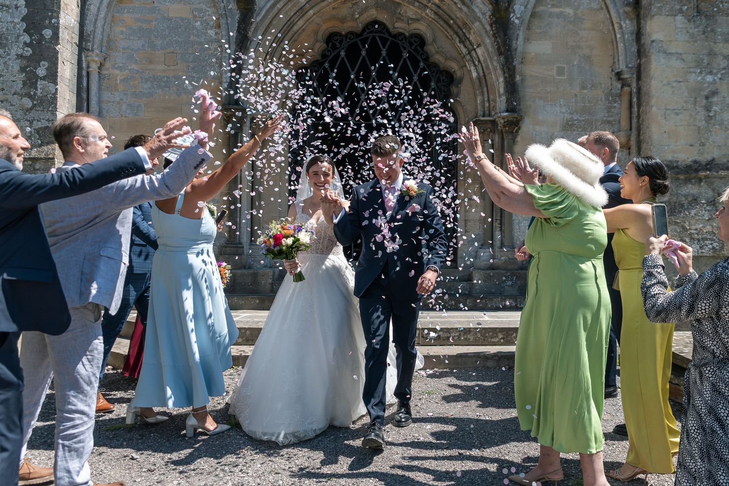 Bride and groom walk through confetti shower outside historic stone church surrounded by smiling wedding guests on sunny day.