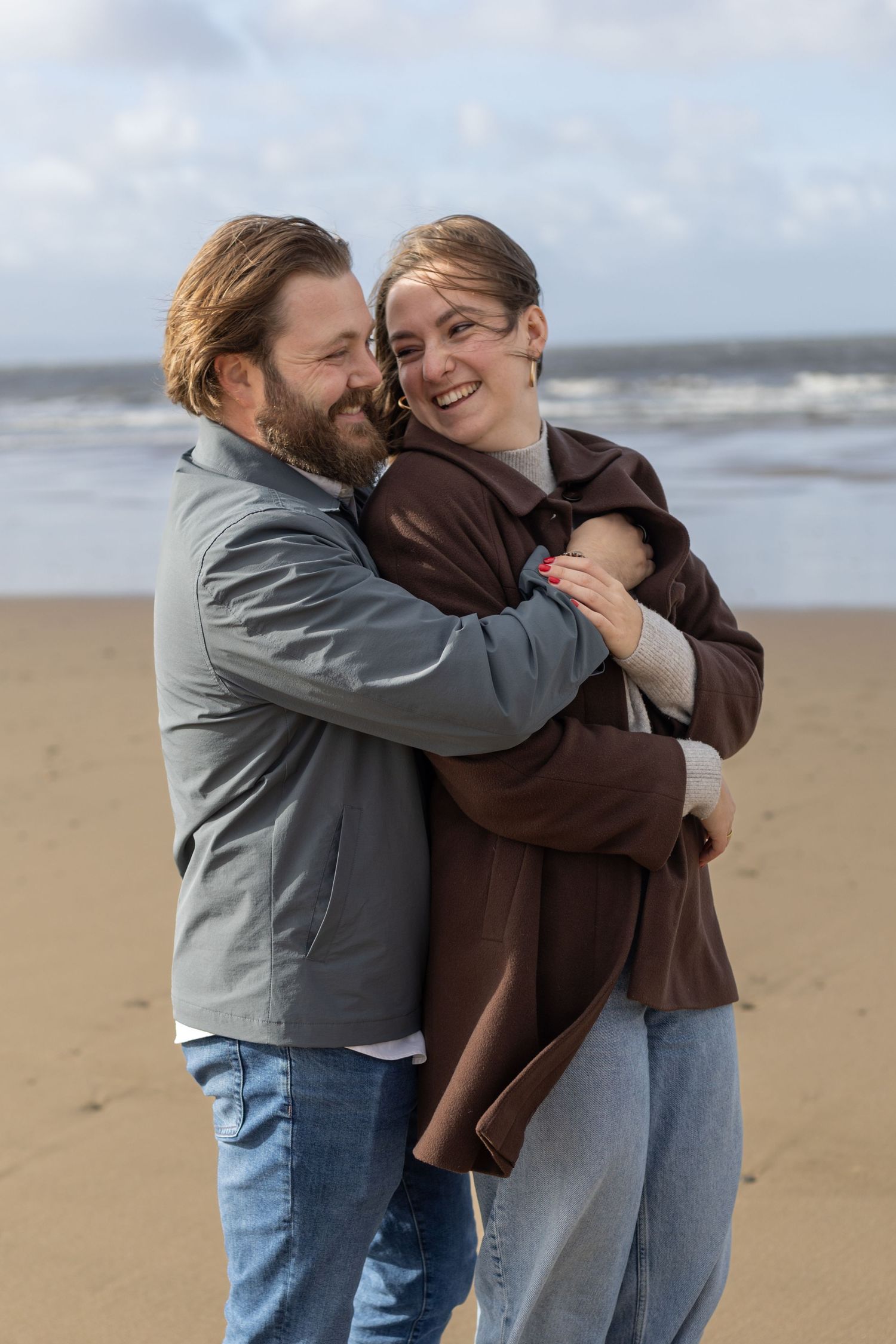 Three people share a group hug while laughing together on a windy beach.