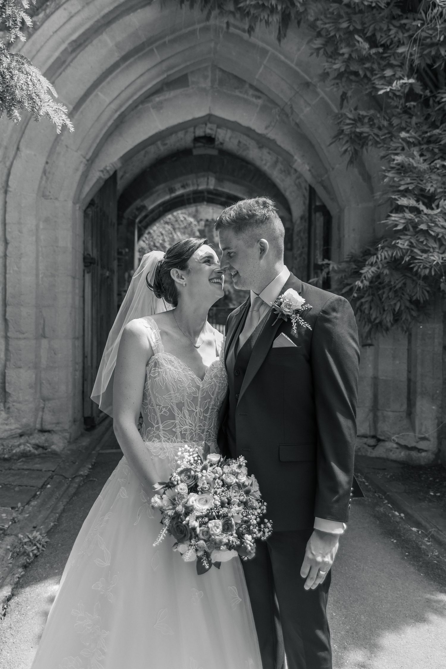 Black and white photo of a bride and groom sharing a romantic moment in front of a stone church archway.
