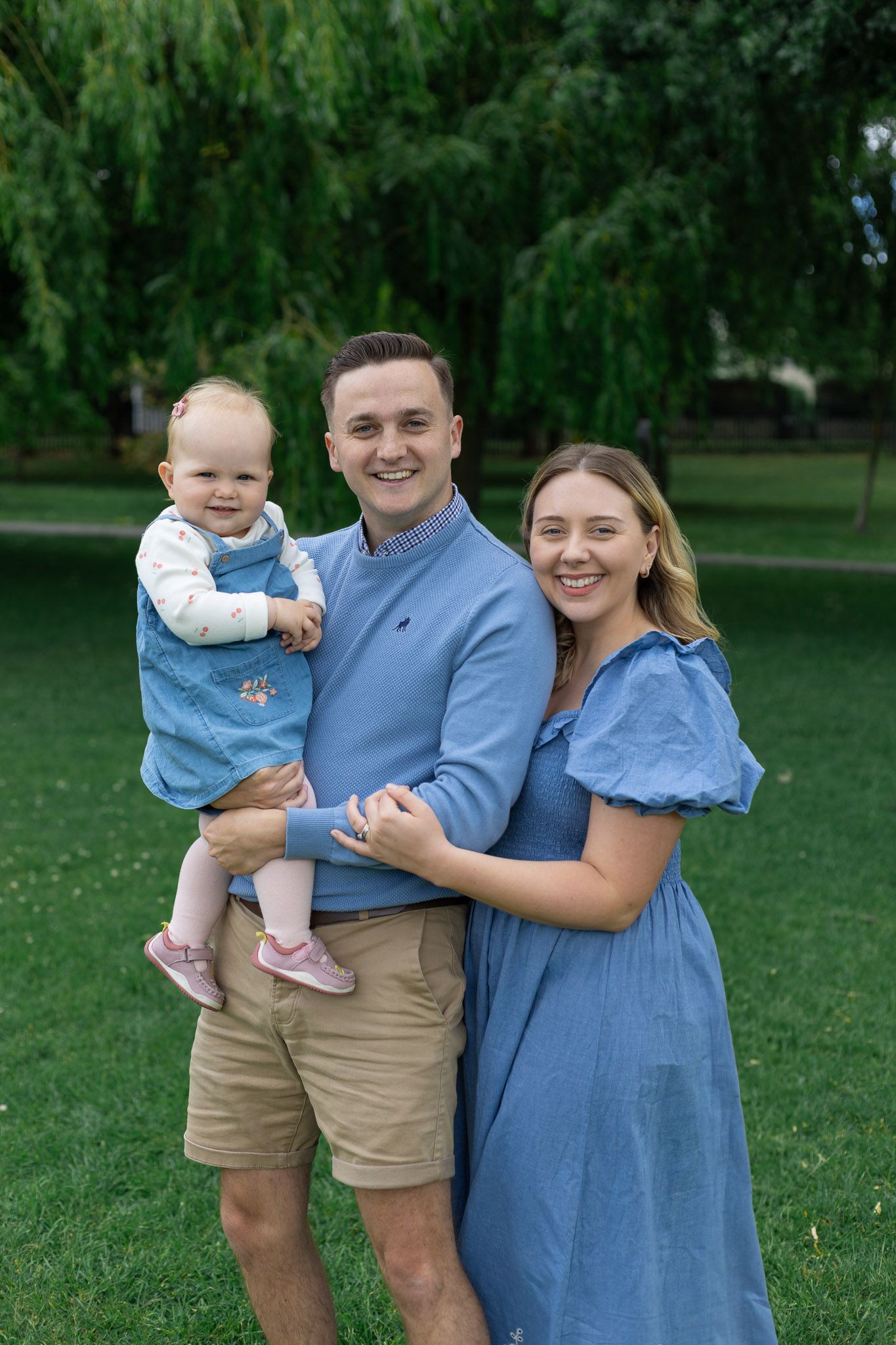 Happy family in matching blue attire embraces while standing on grassy lawn under trees.