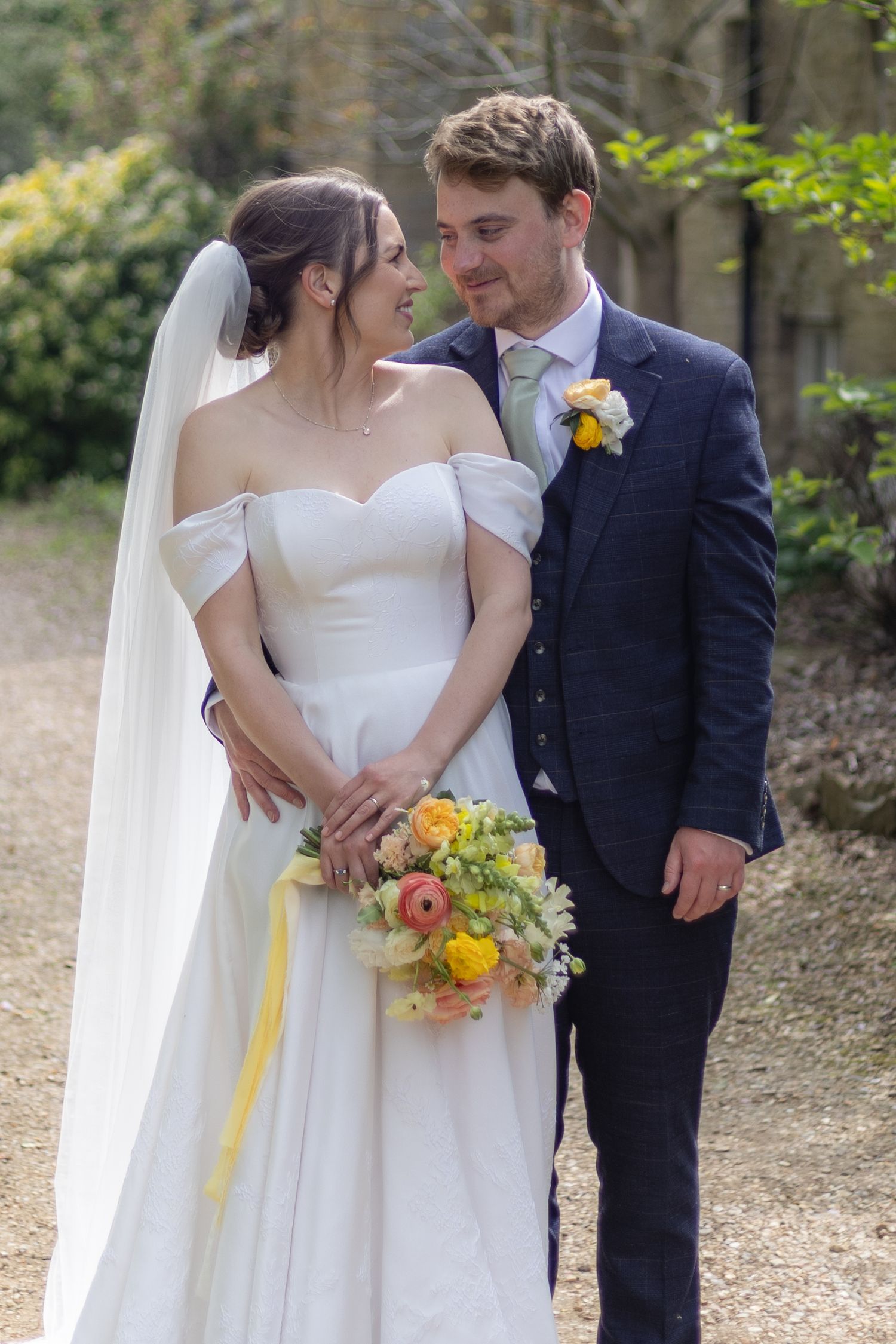 A couple in wedding attire shares intimate moments outdoors while holding a yellow and peach floral bouquet.