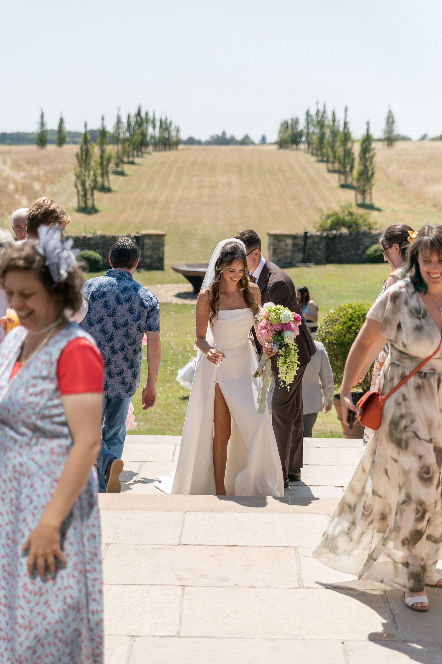 Bride and groom share a kiss outdoors at a sunny wedding venue, surrounded by guests in formal attire.