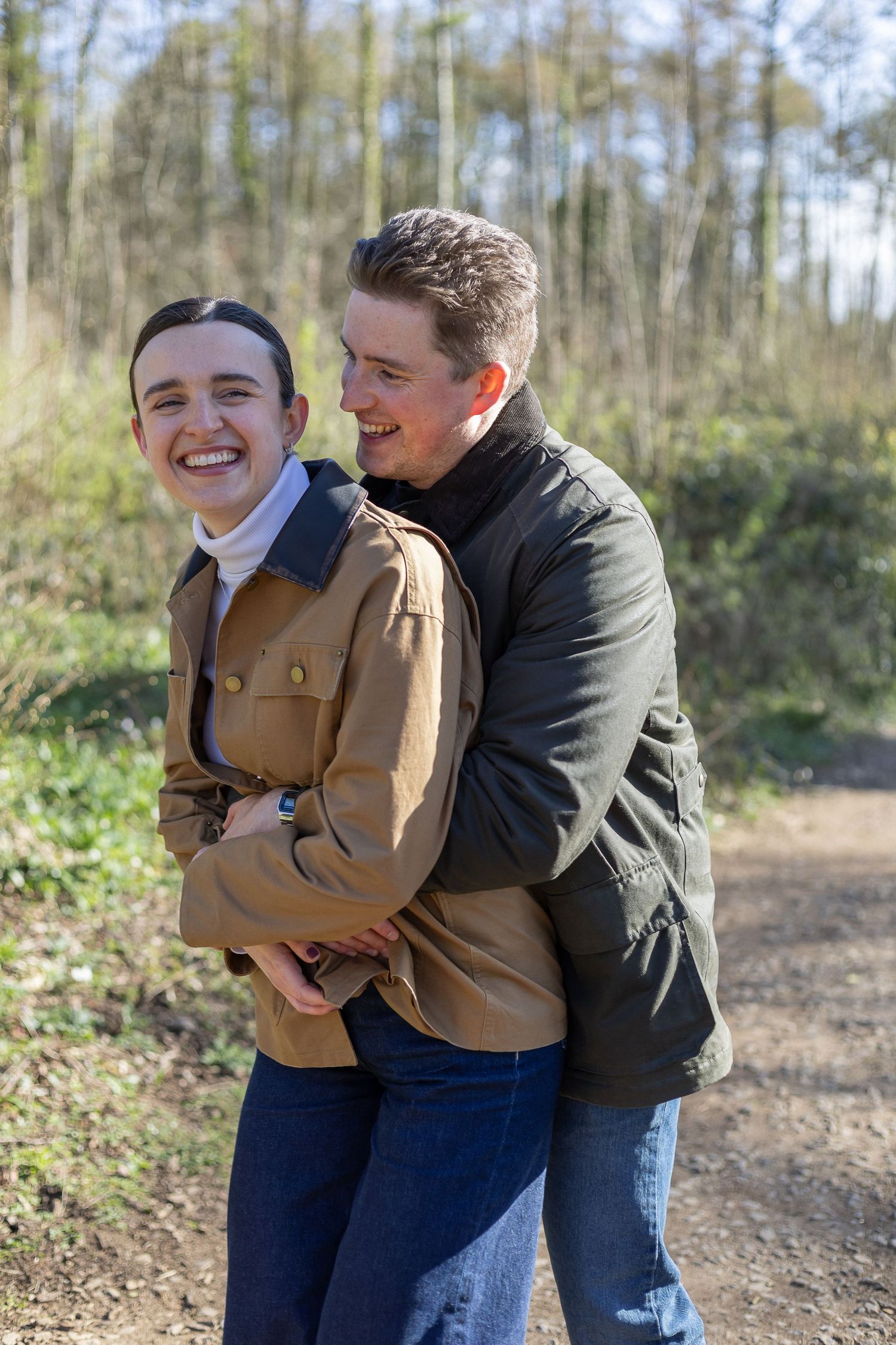 Two people in casual attire share a joyful embrace while standing on a dirt path surrounded by trees.