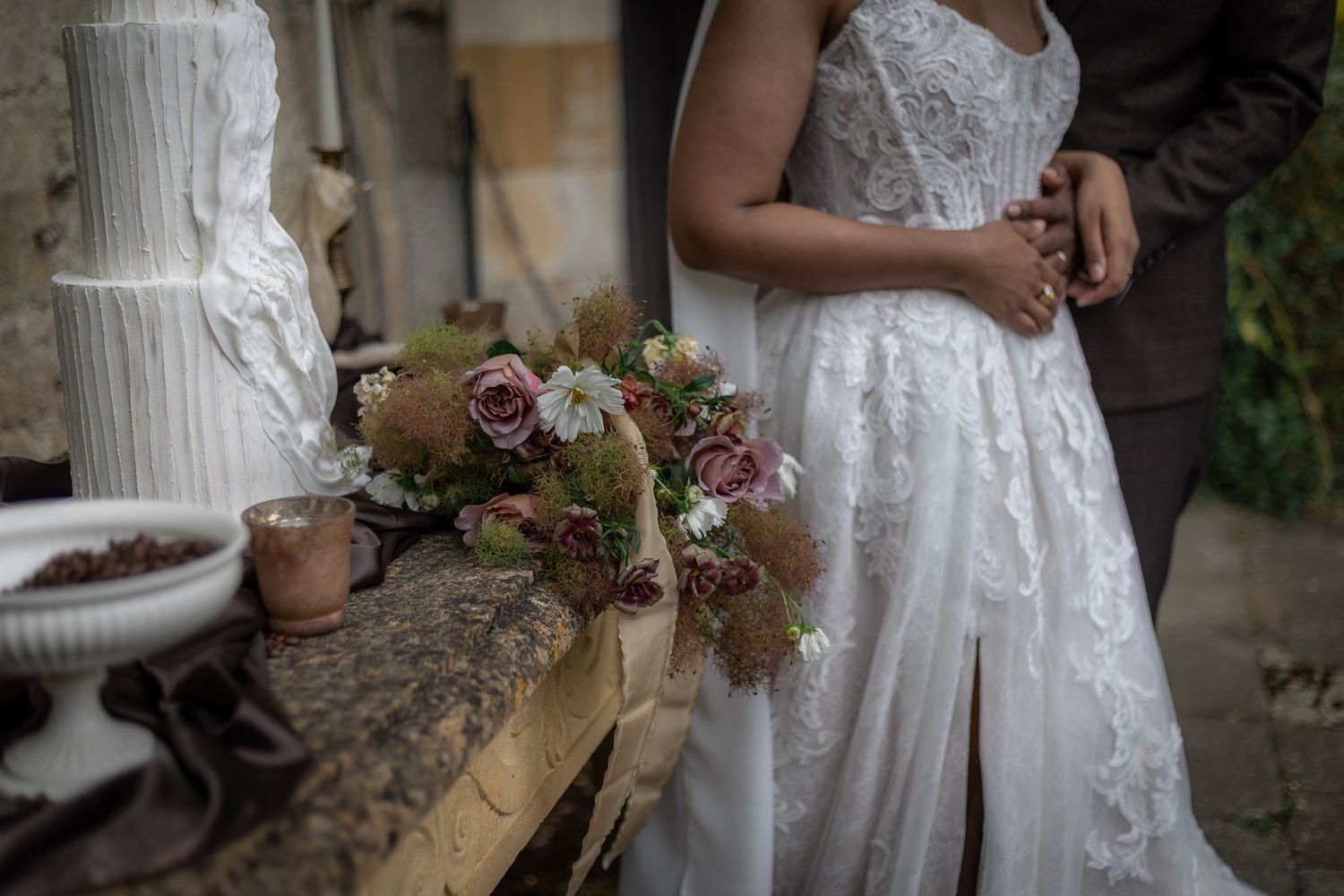 Bride in lace wedding dress holding a bouquet of roses stands beside groom near a rustic wedding cake display at Sudeley Castle