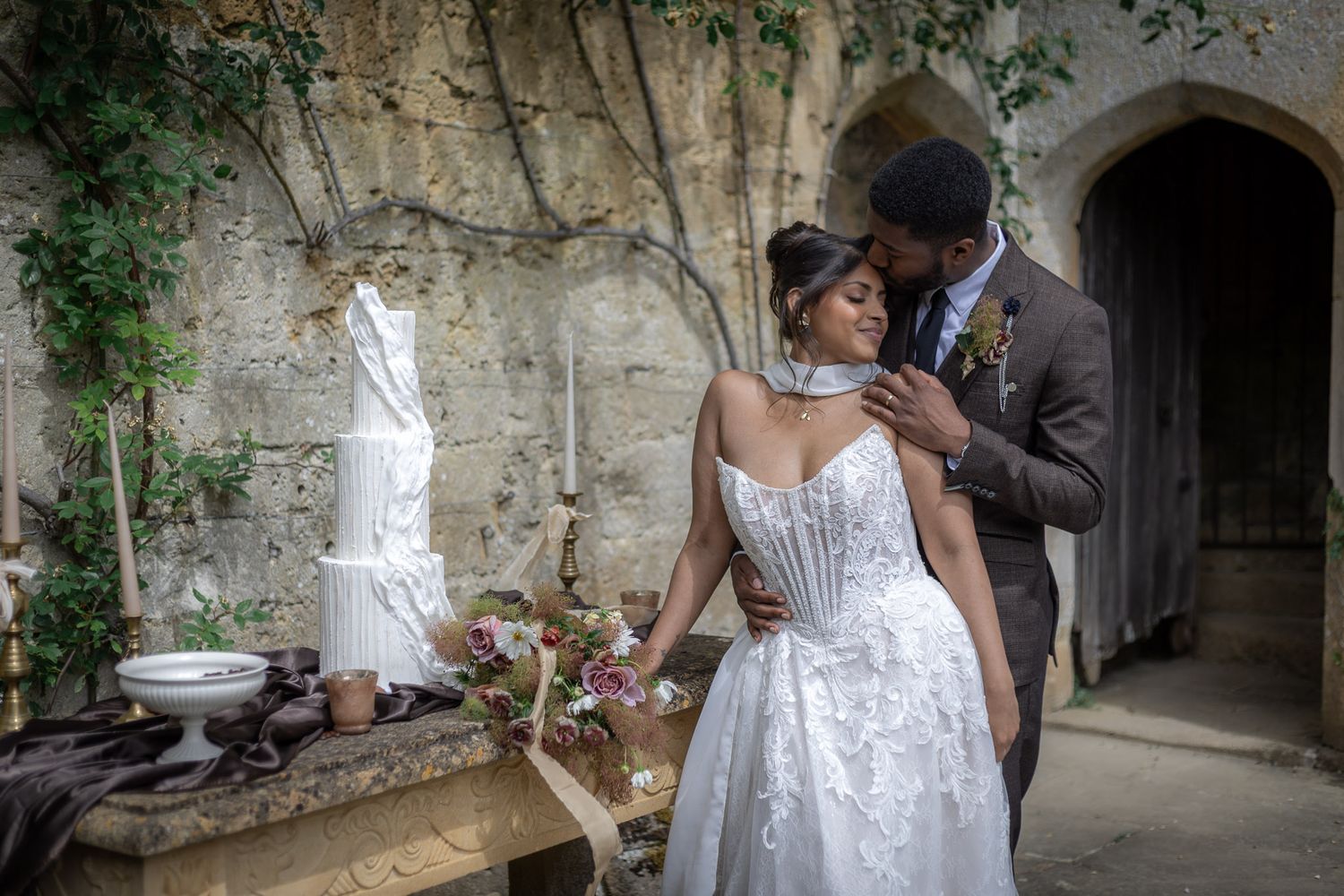 A bride and groom share a romantic moment outdoors near a rustic stone wall with a wedding cake and floral table display at Sudeley Castle