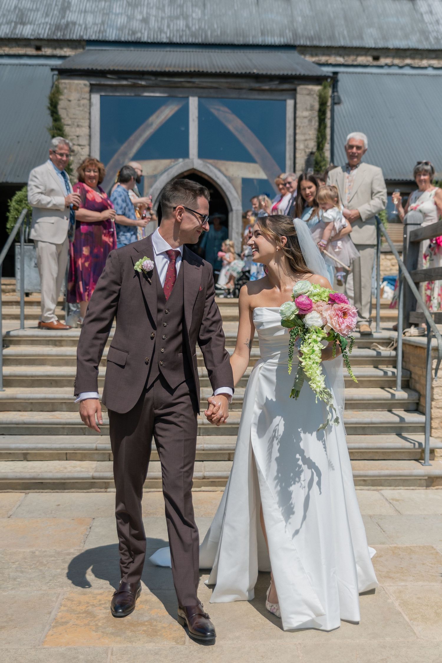 Bride and groom hold hands walking from church steps, guests celebrating behind them, bride holding pink bouquet.