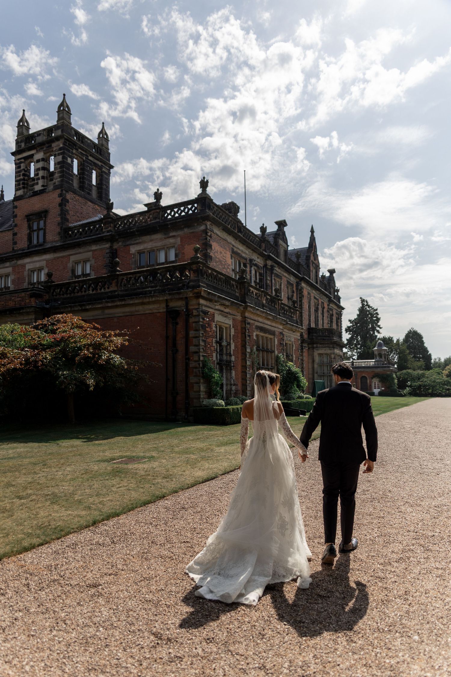 A couple walks along a gravel path leading to a grand Victorian Gothic mansion on a cloudy summer day.