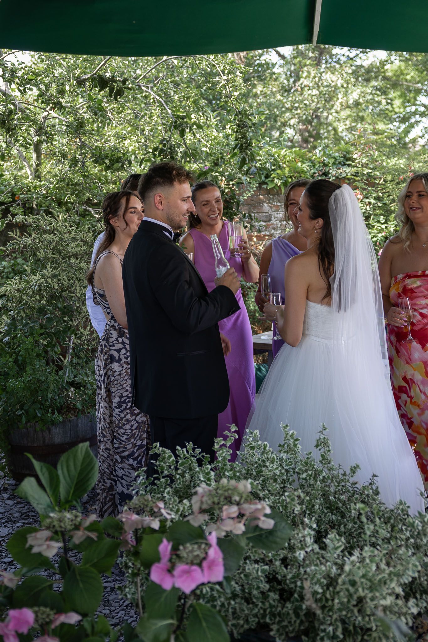Wedding party gathers under green tent surrounded by garden flowers.