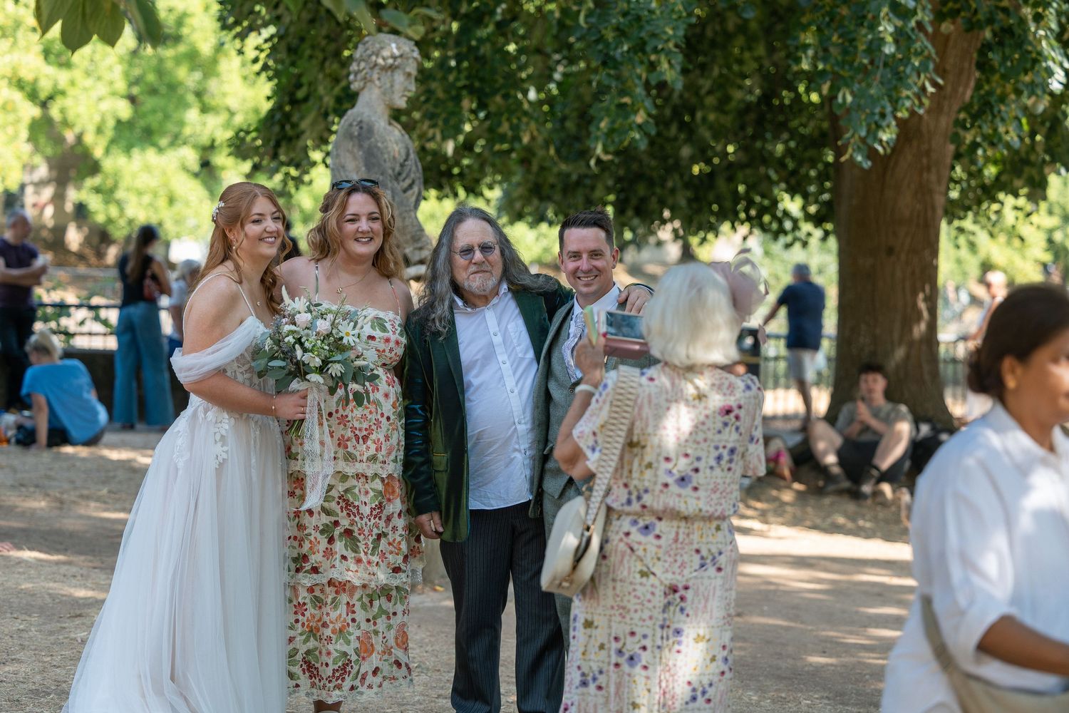 Two brides in white dresses pose with guests outdoors at a garden wedding ceremony under lush green trees in Parade Gardens, Bath