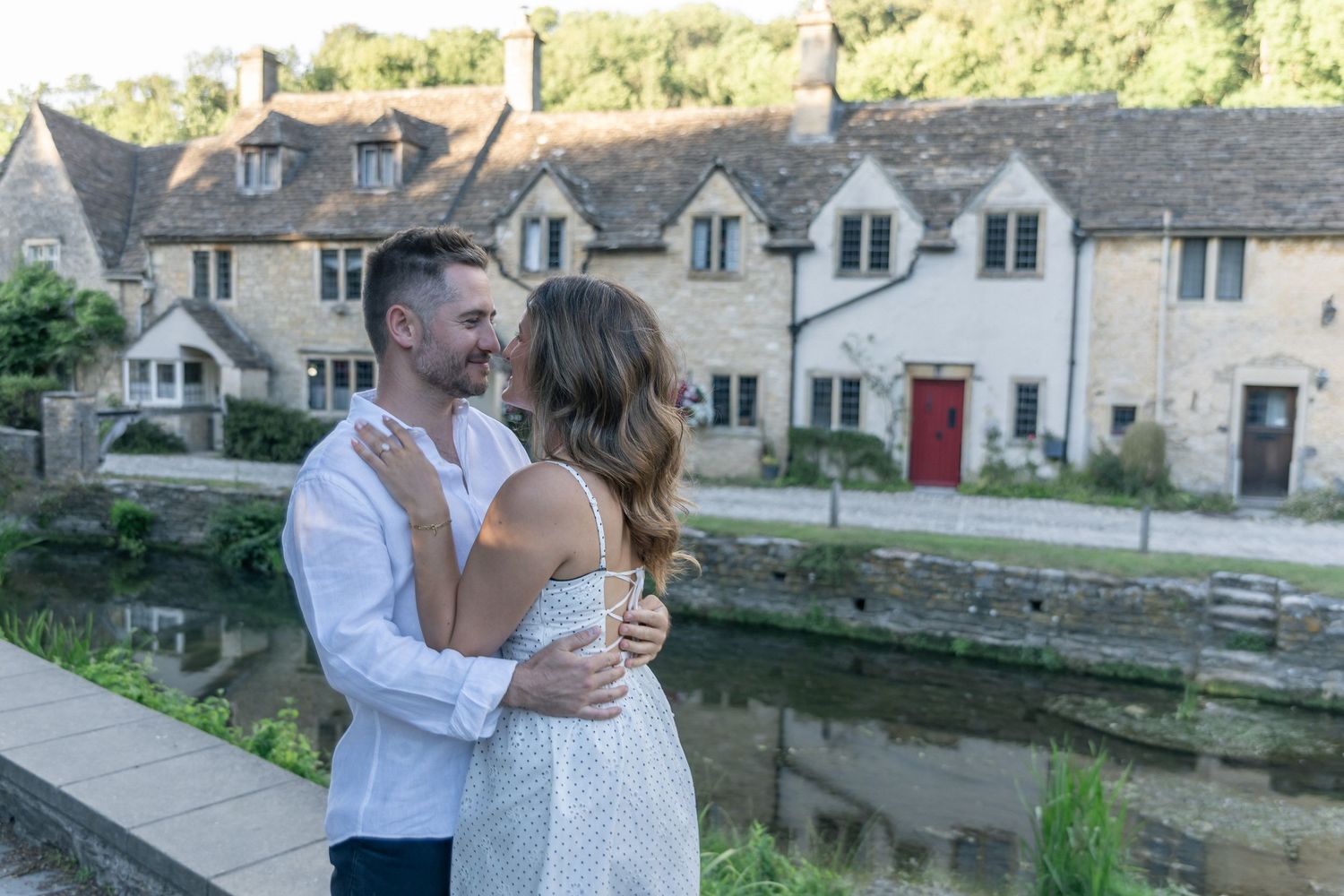 Couple embracing and smiling by a tranquil river with charming Cotswolds stone cottages in the background.