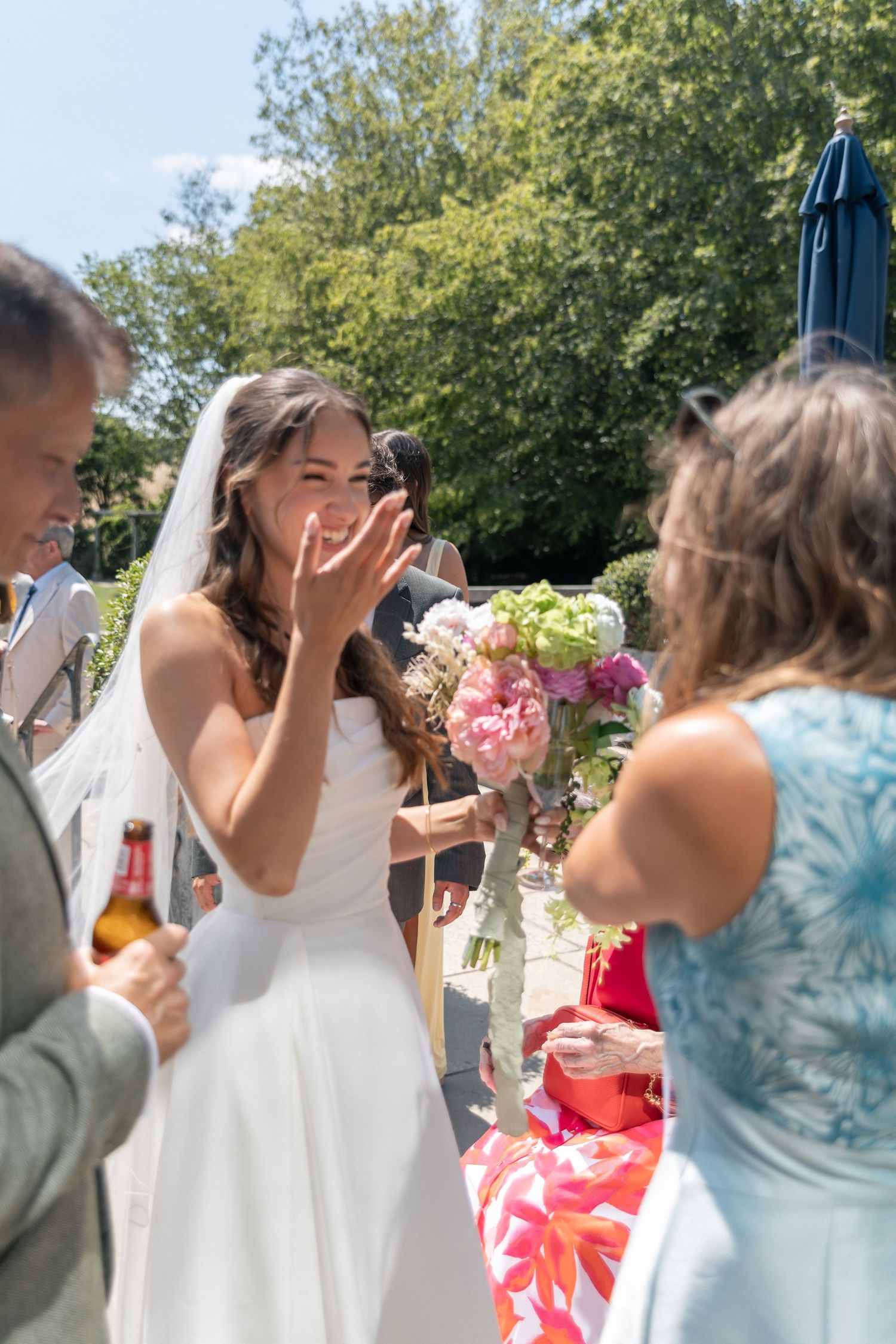 Bride in white gown and veil waves to guests while holding pink bouquet at sunny outdoor wedding reception.