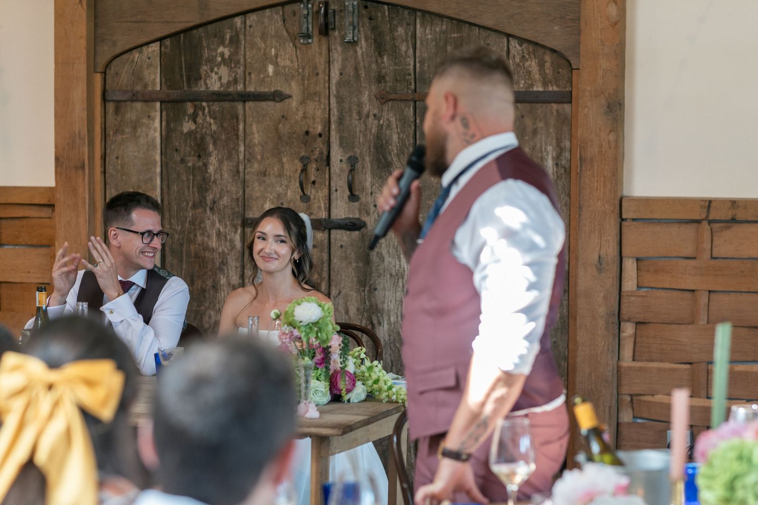 Bride and groom smile during wedding reception speech at rustic barn venue with wooden doors.
