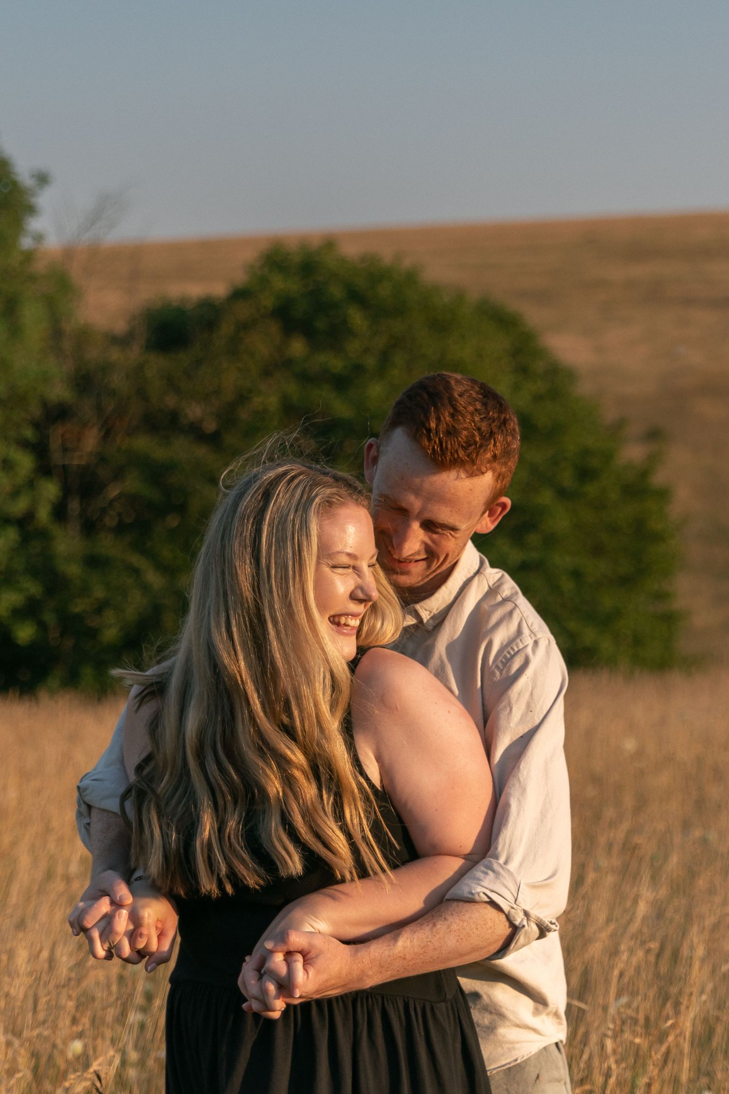A couple embraces in a golden field at sunset, the man hugging the woman from behind as they smile joyfully.