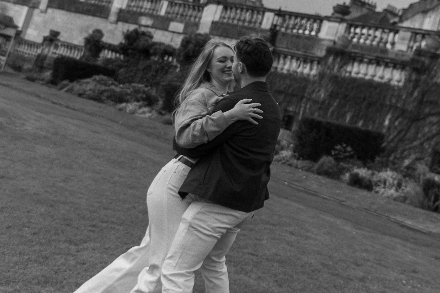 Couple dancing outdoors on grass in black and white photo with historic building in background.