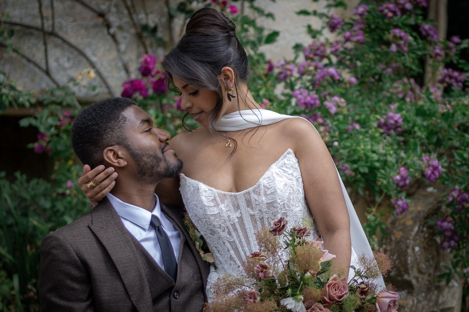 Groom in brown suit kisses bride in white lace gown holding pink bouquet, surrounded by purple flowering vines at Sudeley Castle