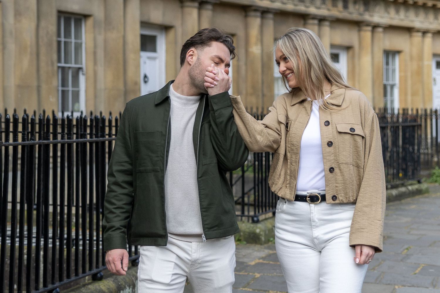 A young couple walking outdoors, woman in beige jacket comforting laughing man in green jacket on a city street.