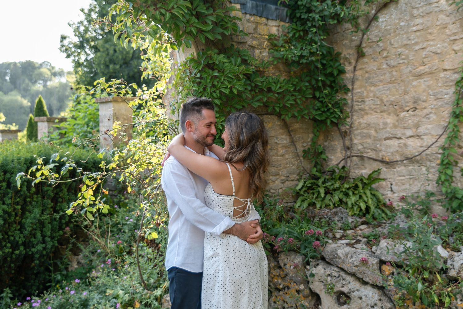 Couple embracing and smiling near a stone wall covered in ivy, surrounded by lush greenery in a romantic outdoor setting.