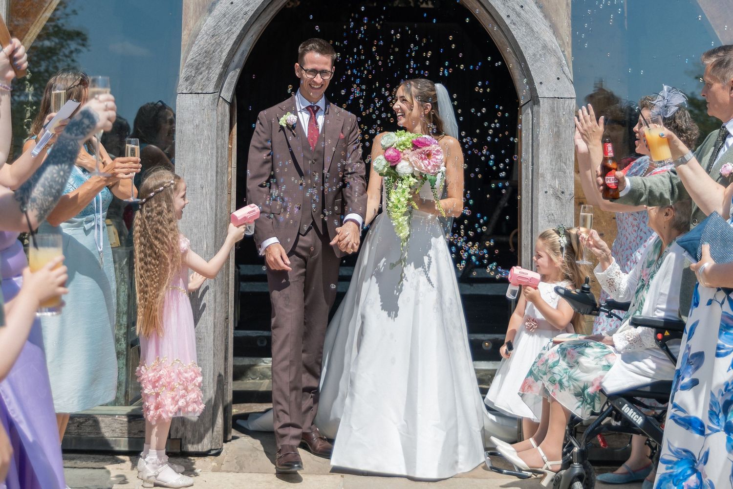 Newlywed couple exits church amid confetti shower, surrounded by cheering guests on a sunny wedding day.