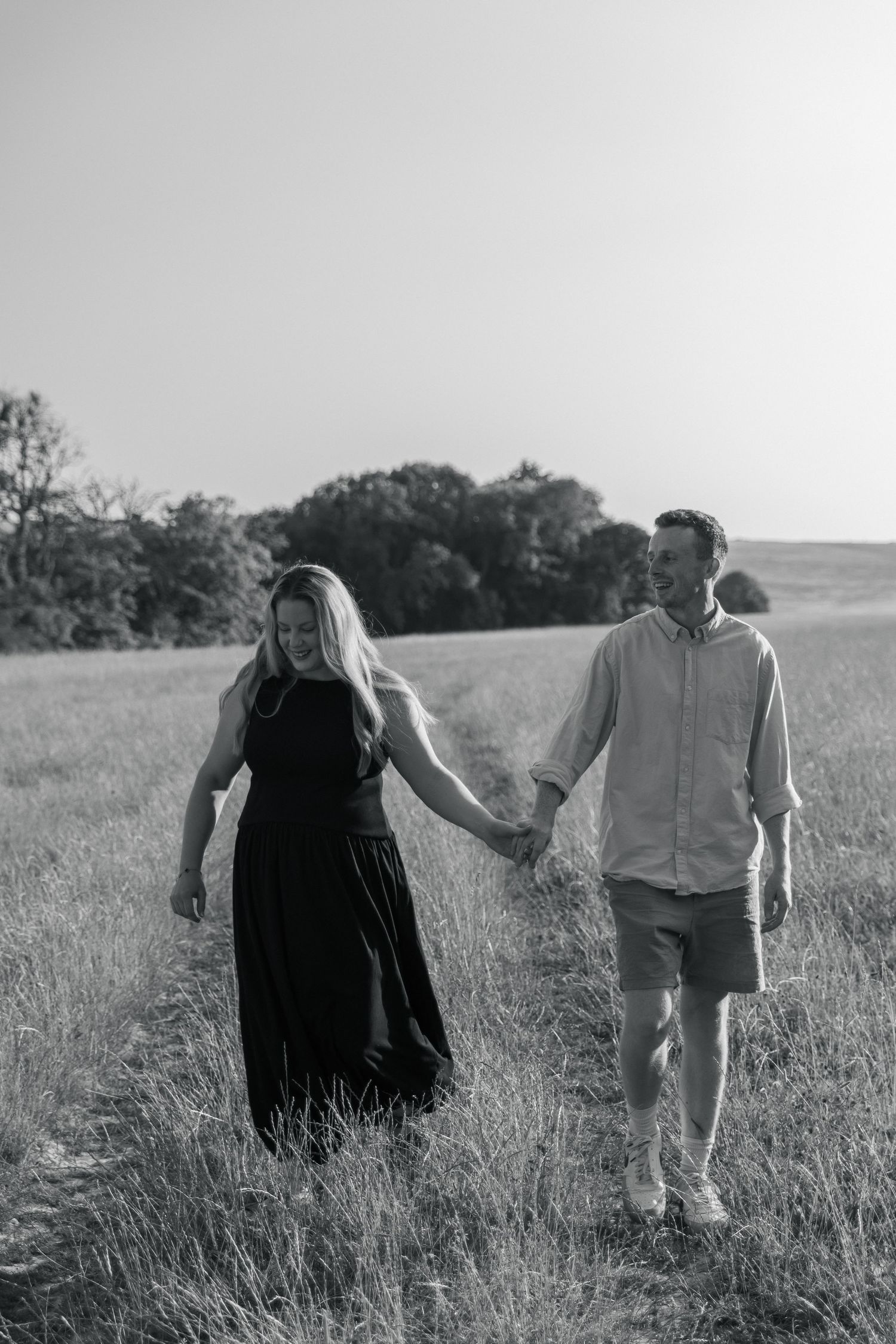 Black and white photo of a couple holding hands walking through a grassy field with trees in the background.