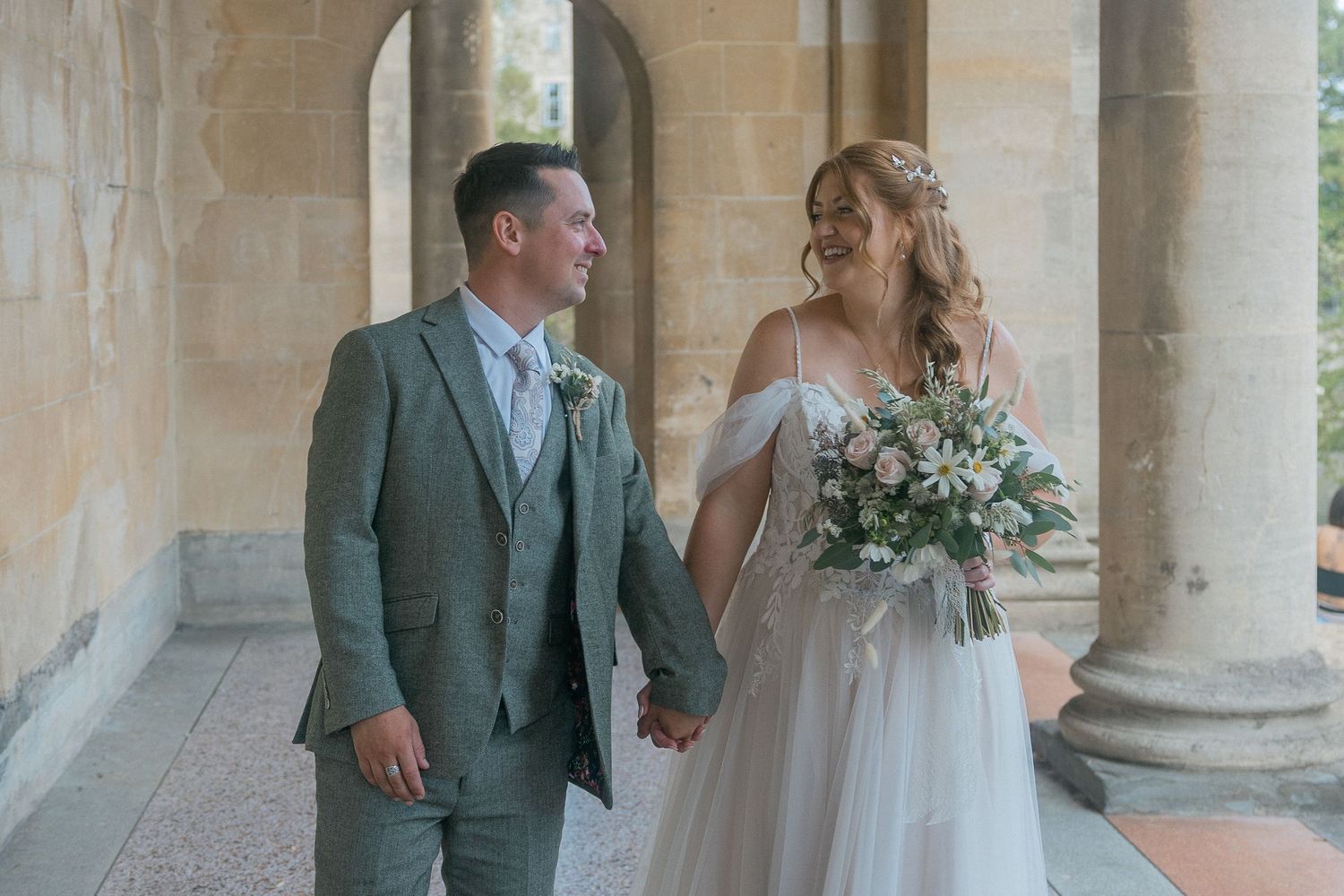 A bride and groom hold hands and smile at each other in a sunlit stone colonnade, she holds a pastel bouquet.