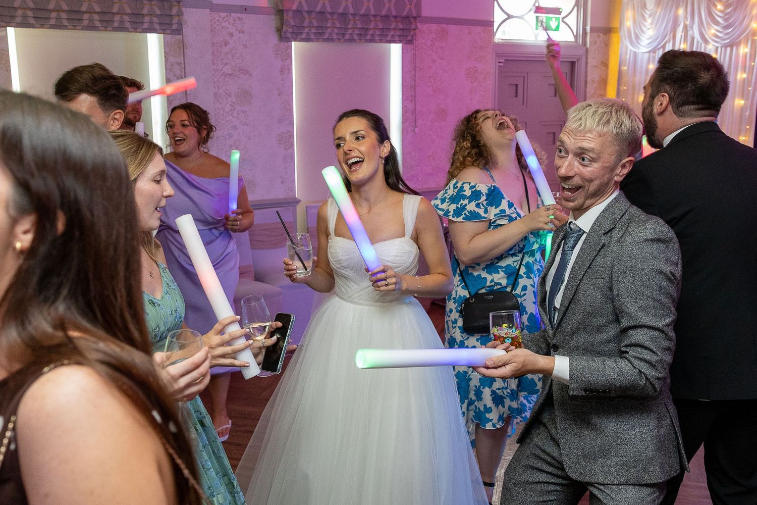 Wedding guests dance and celebrate together on a purple-lit dance floor during a reception party.