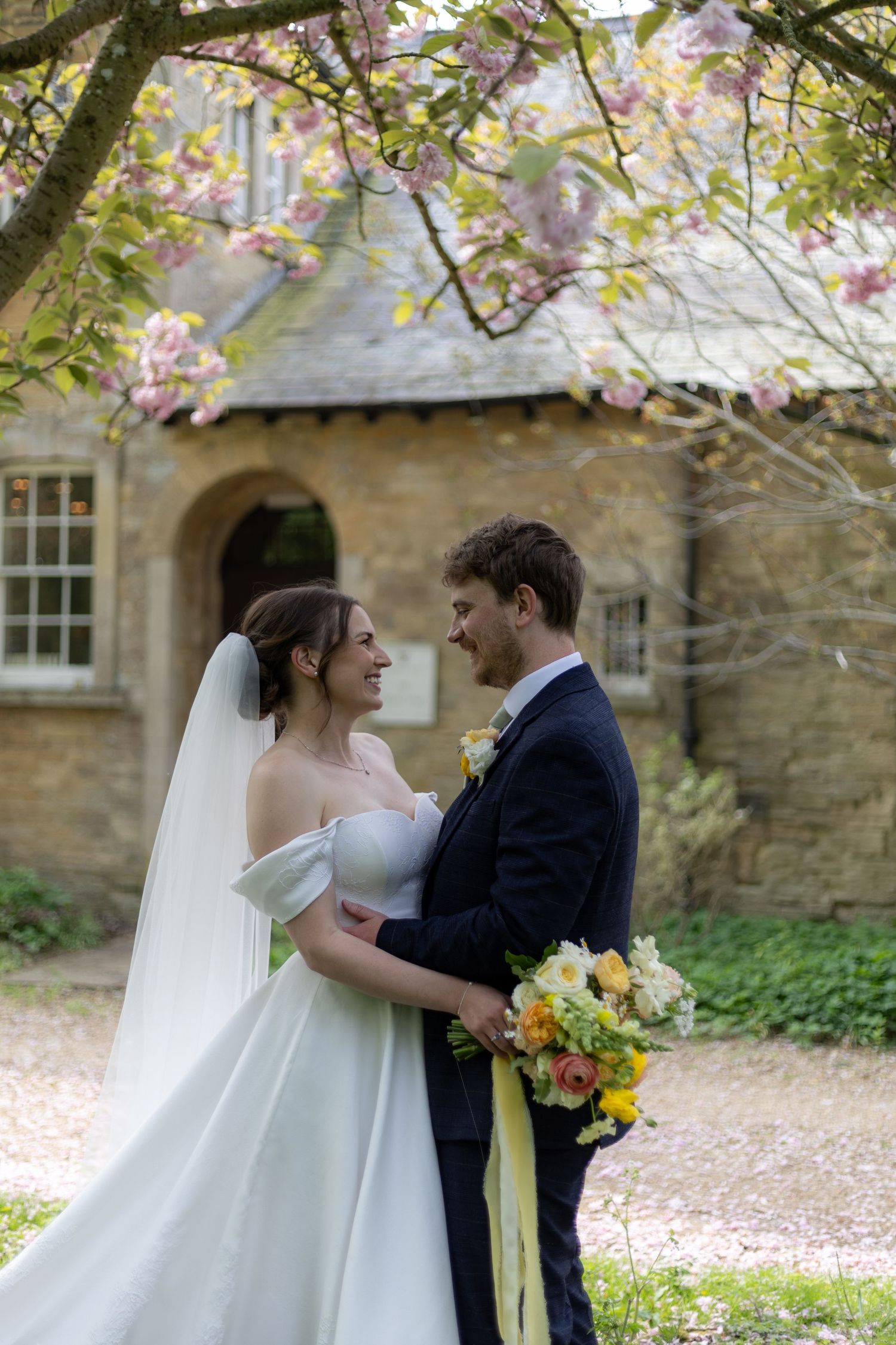 A romantic wedding couple shares intimate moments under blooming spring trees at a rustic stone venue.