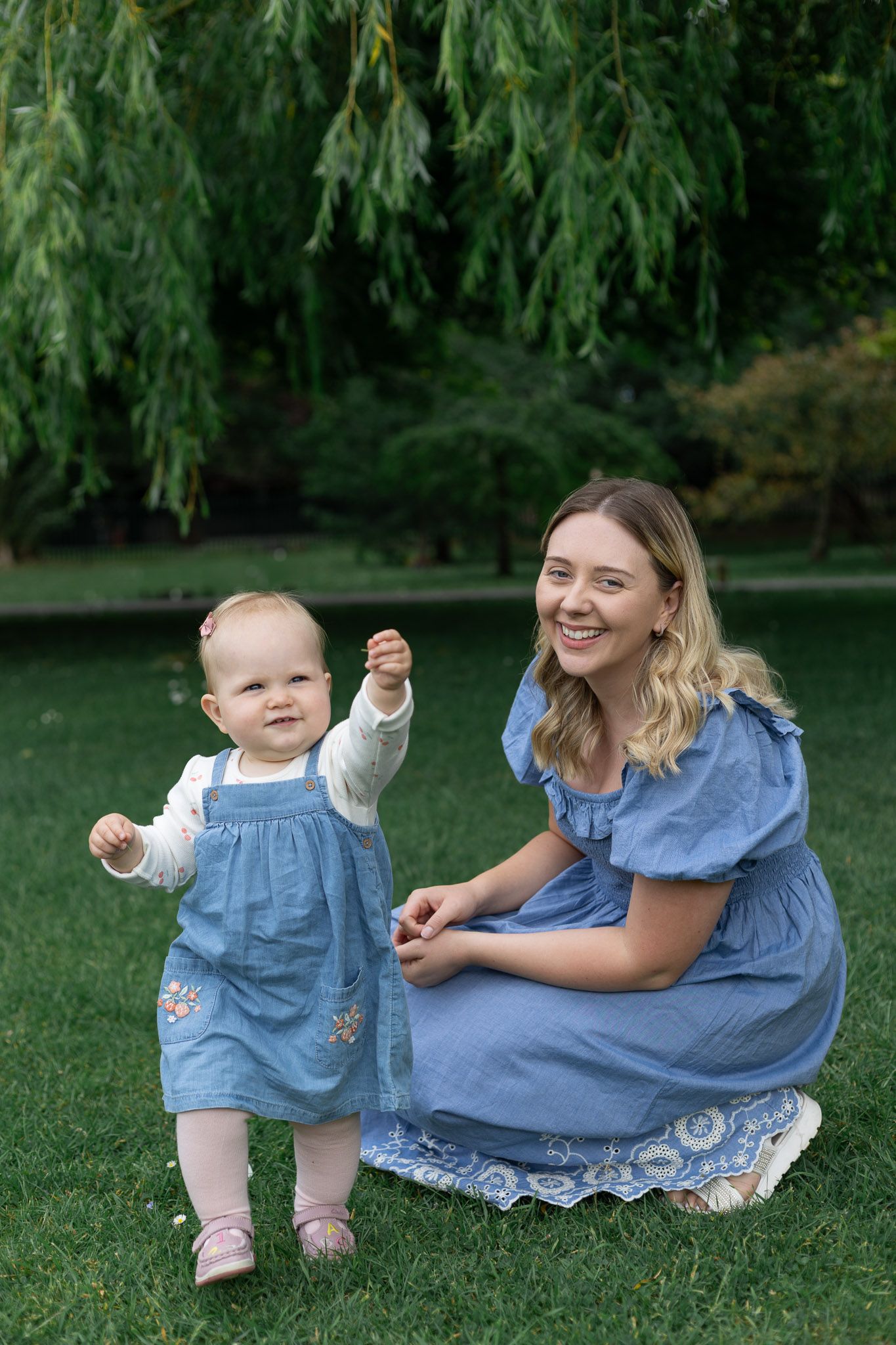 Mother and baby in matching blue dresses play together on grass under willow trees.