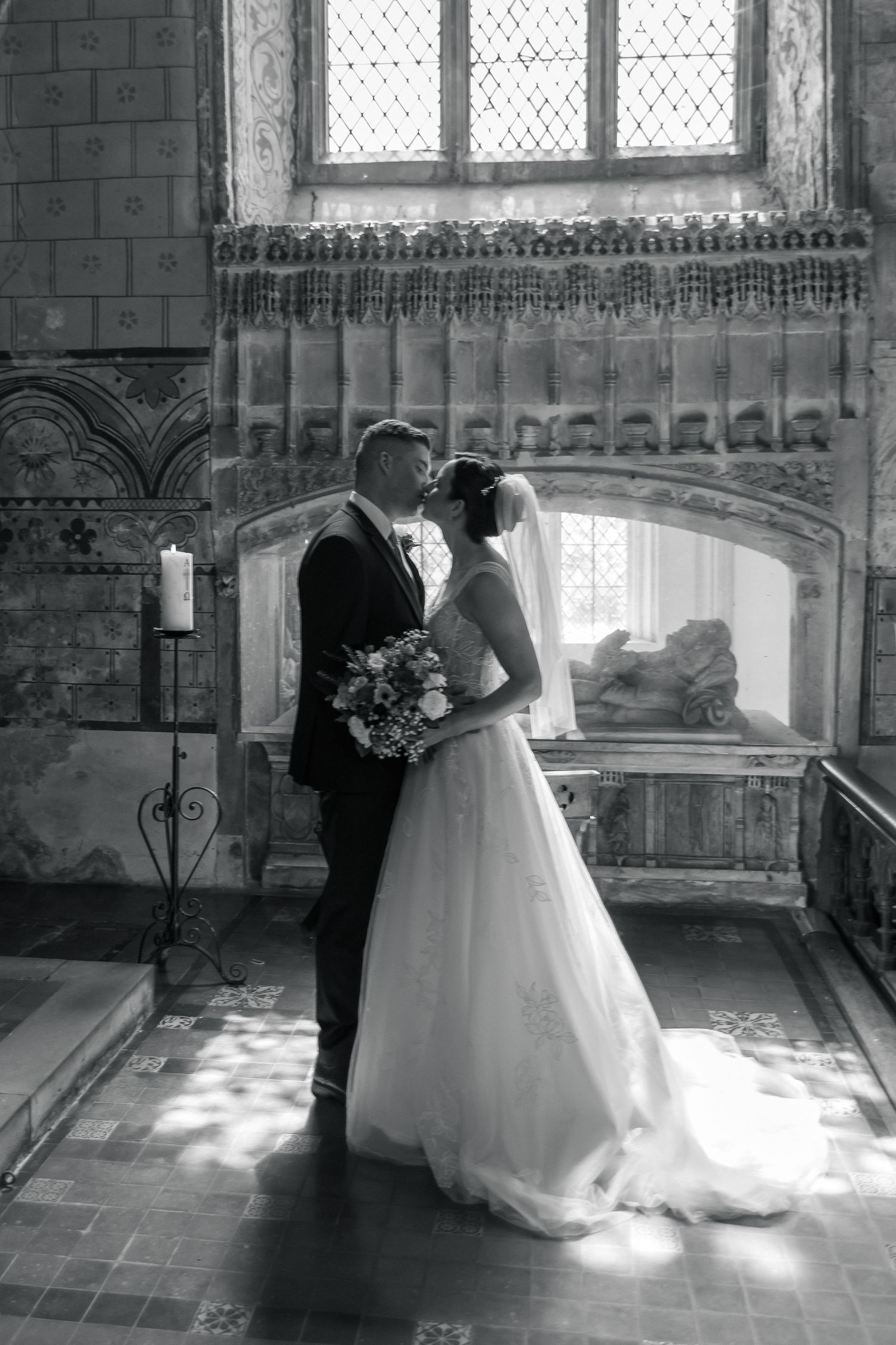 Black and white photo of bride and groom kissing in a historic church, bride holding bouquet in elegant wedding gown.