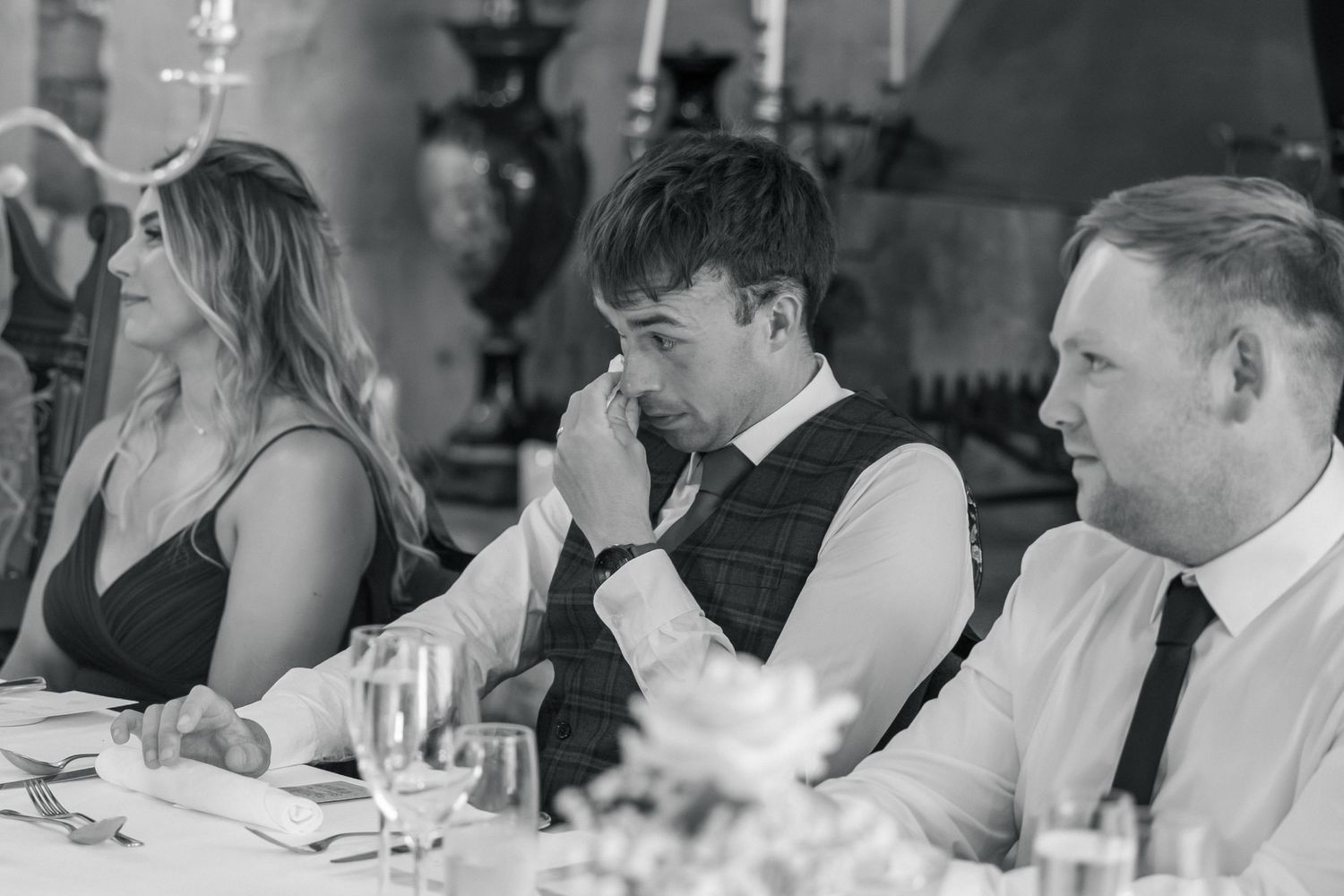 Emotional wedding guest wiping tears at reception table, seated between two others in black and white photo.