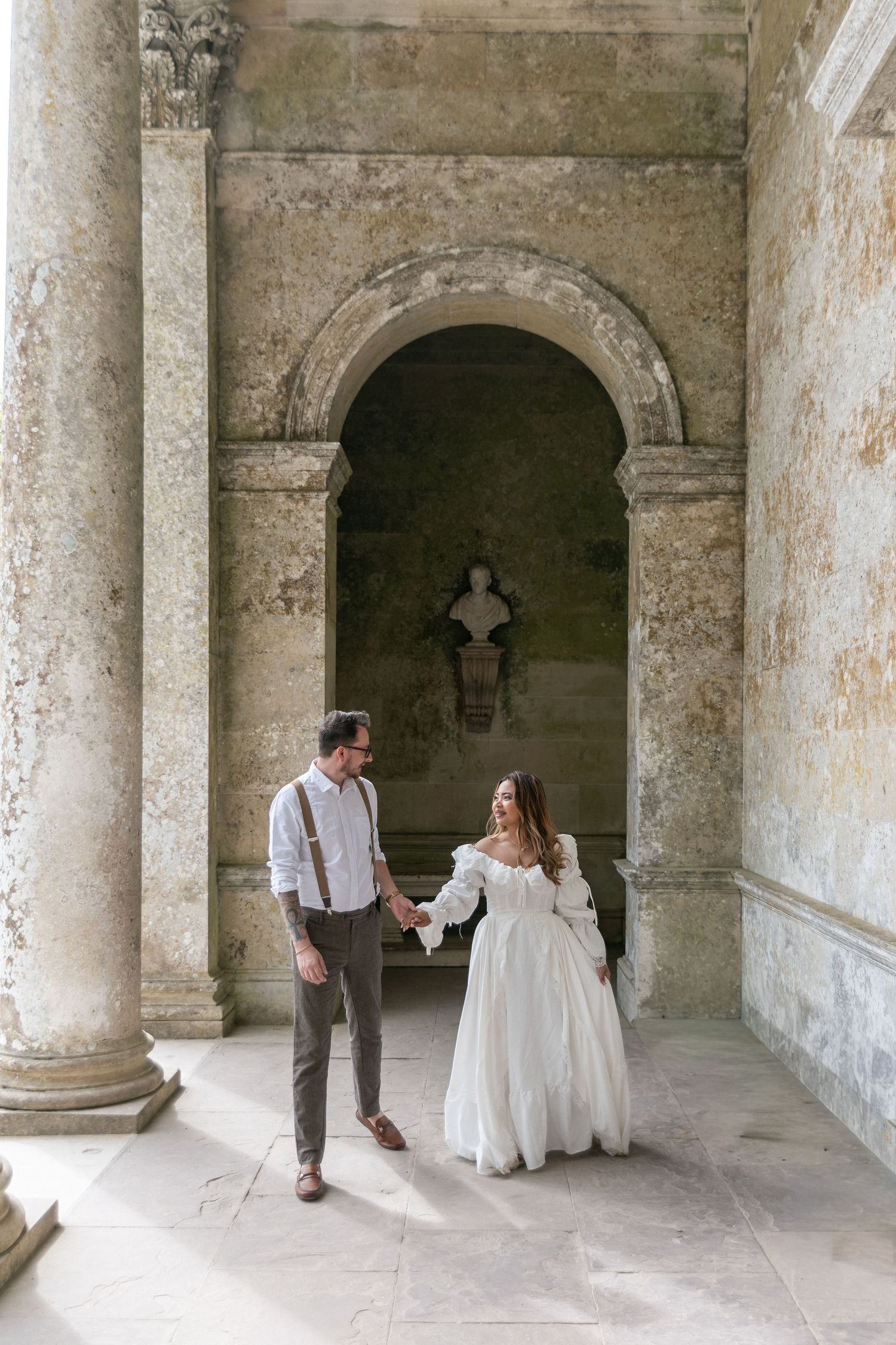 Couple in wedding attire holding hands in a historic stone archway with classical columns and a bust sculpture.