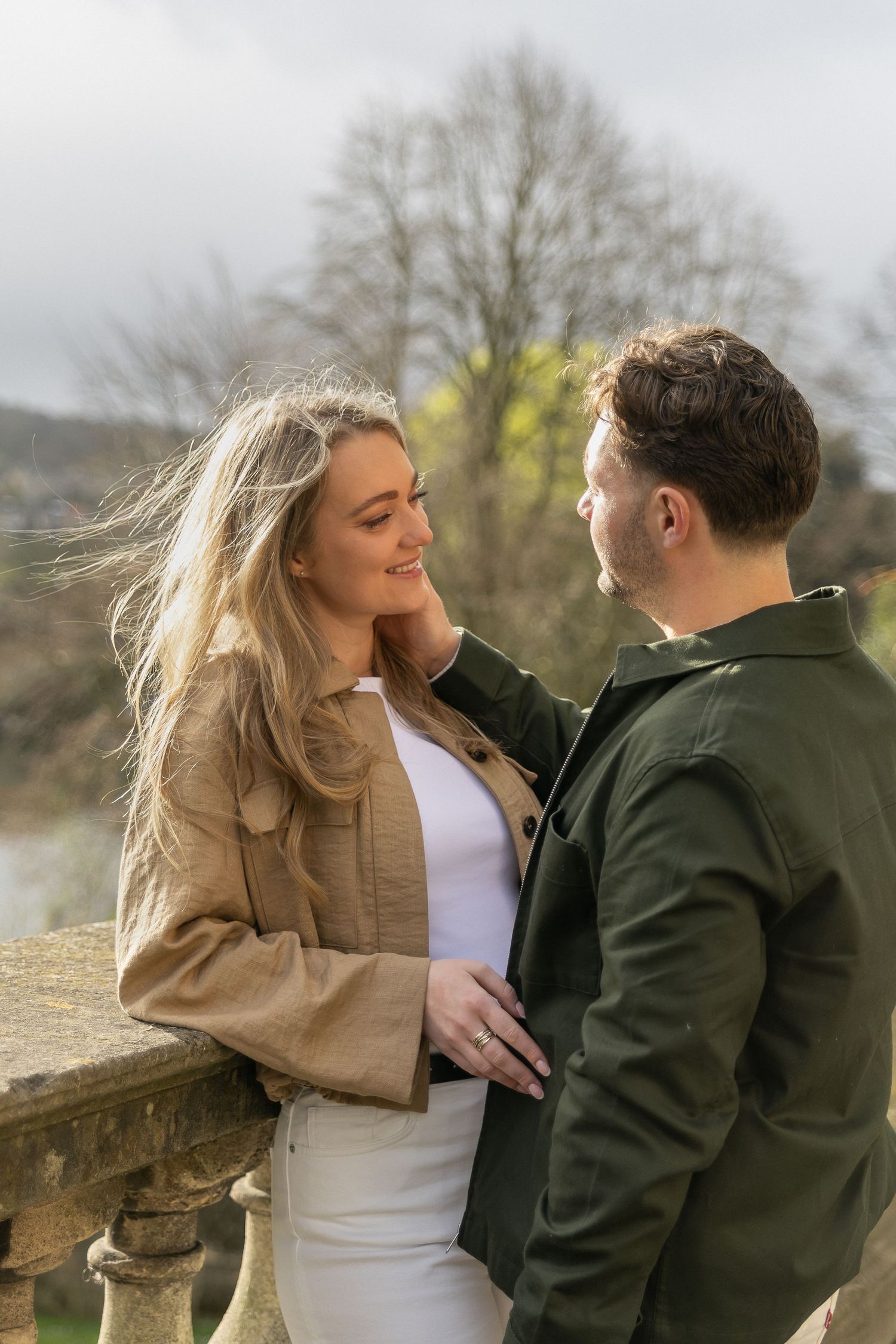 A smiling couple sharing a romantic moment outdoors, woman in beige jacket touching mans face, wearing engagement ring.