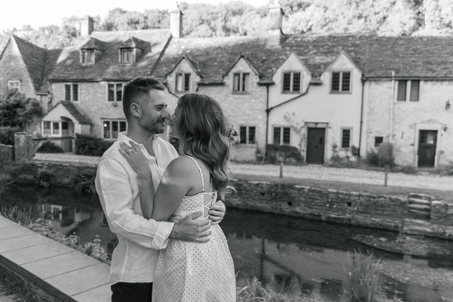 Black and white photo of a couple embracing and smiling by a canal with charming stone cottages in the background.
