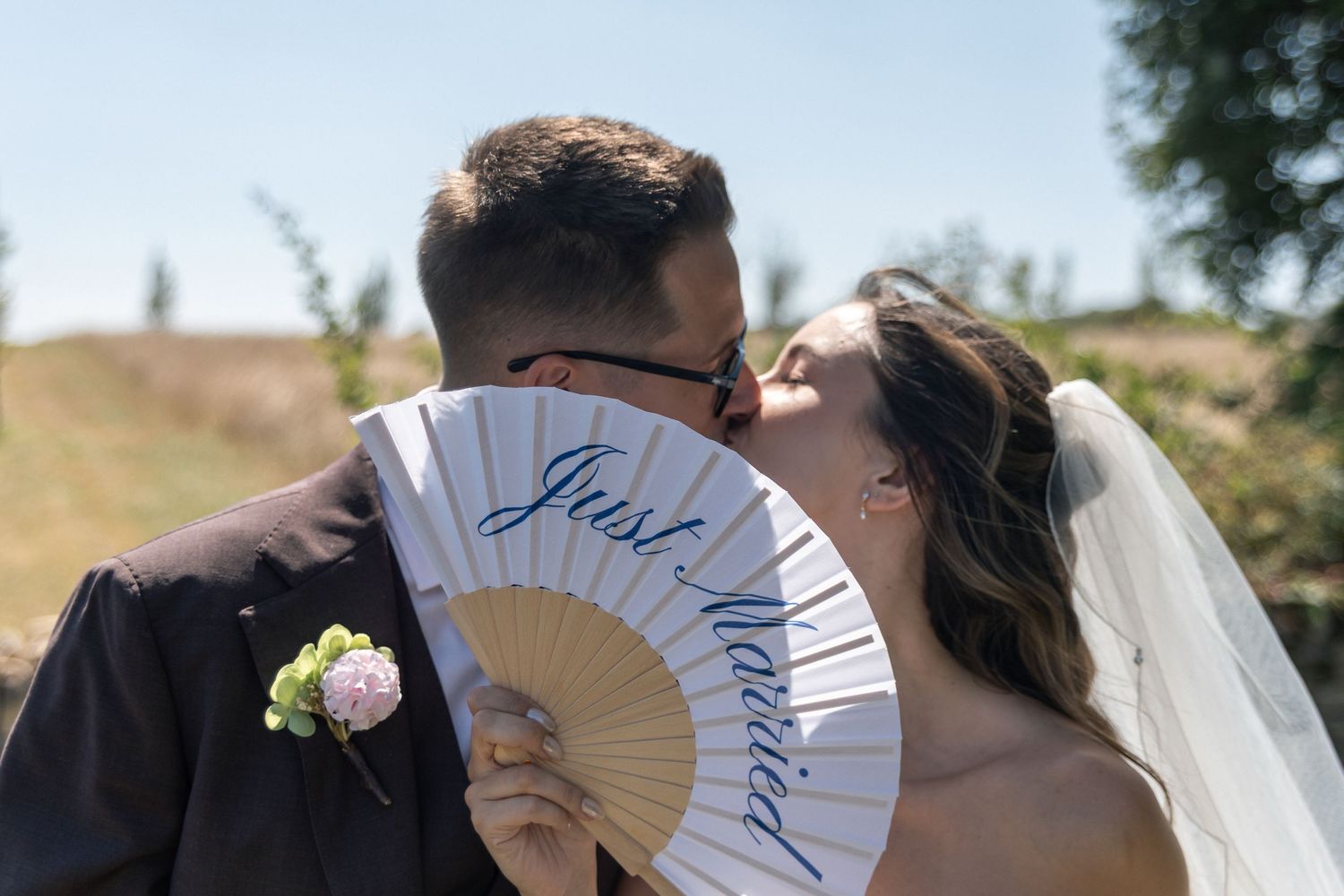 Newlywed couple kissing behind a decorative hand fan reading Just Married on their wedding day outdoors.