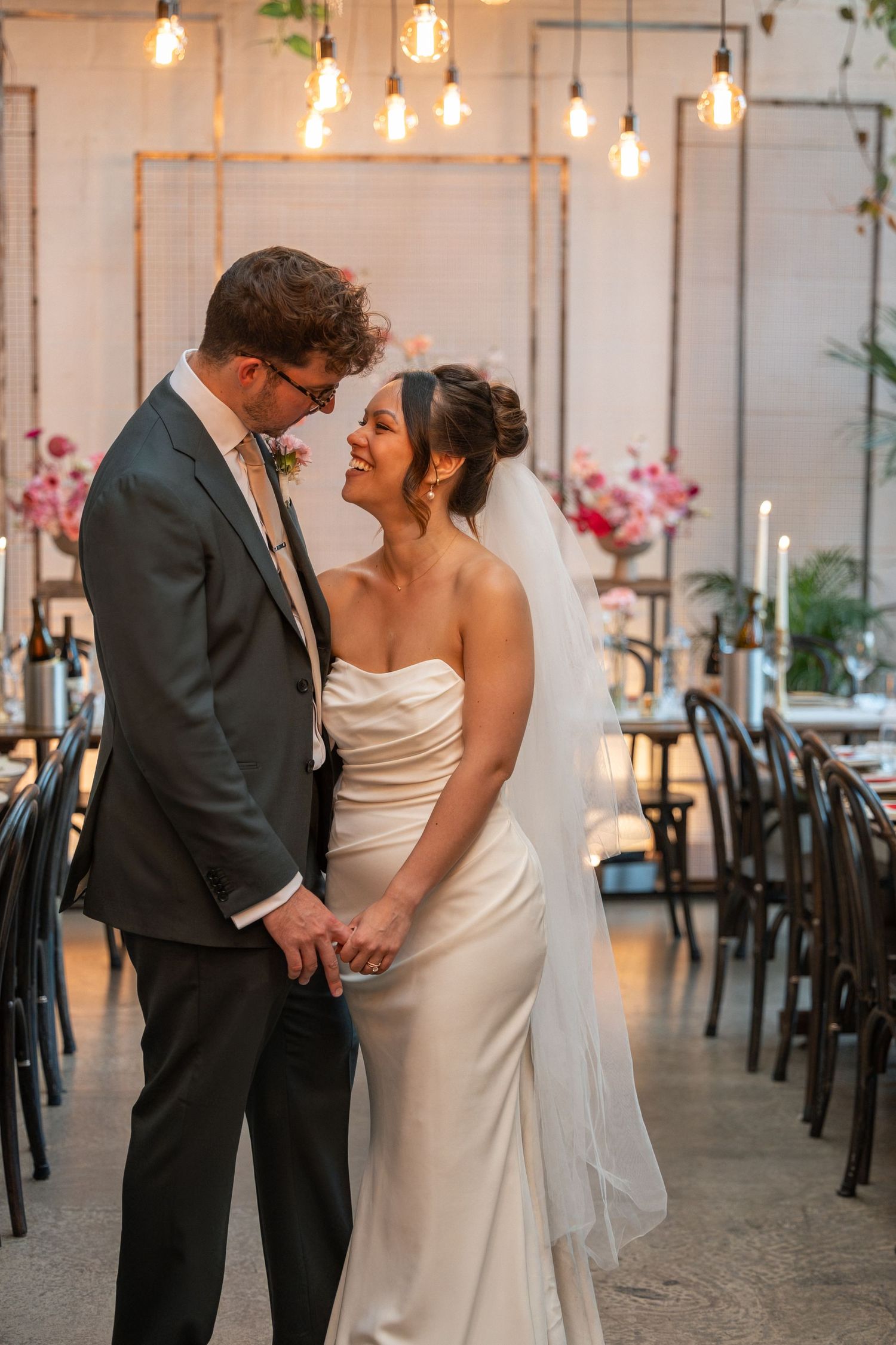 A bride and groom share a joyful moment holding hands in an elegantly decorated wedding reception venue with warm Edison bulb lighting.