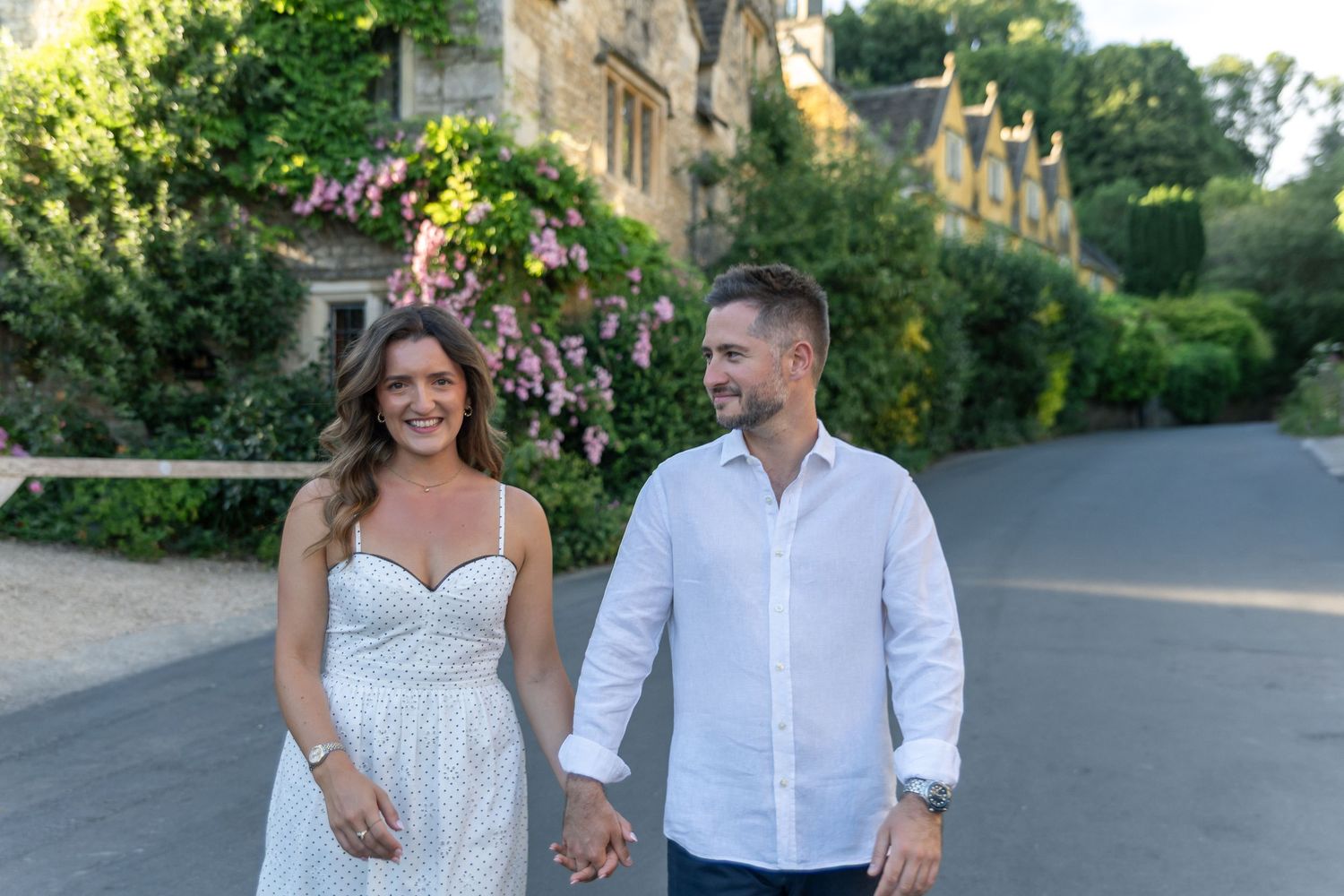 Couple holding hands smiling while walking on a sunlit path beside a stone building with lush greenery and flowers.
