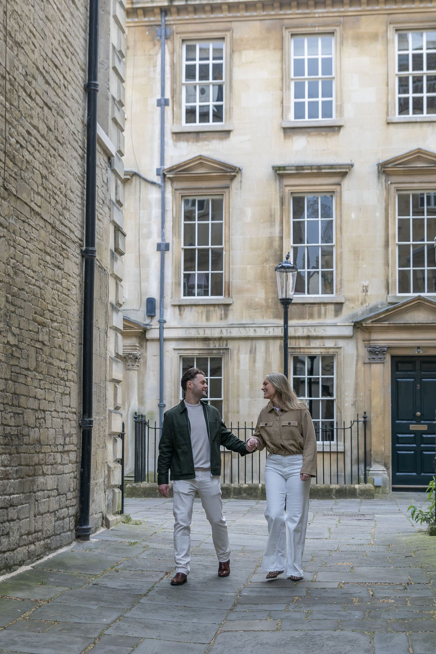 Couple holding hands walking through a charming cobblestone alley beside a Georgian stone building.