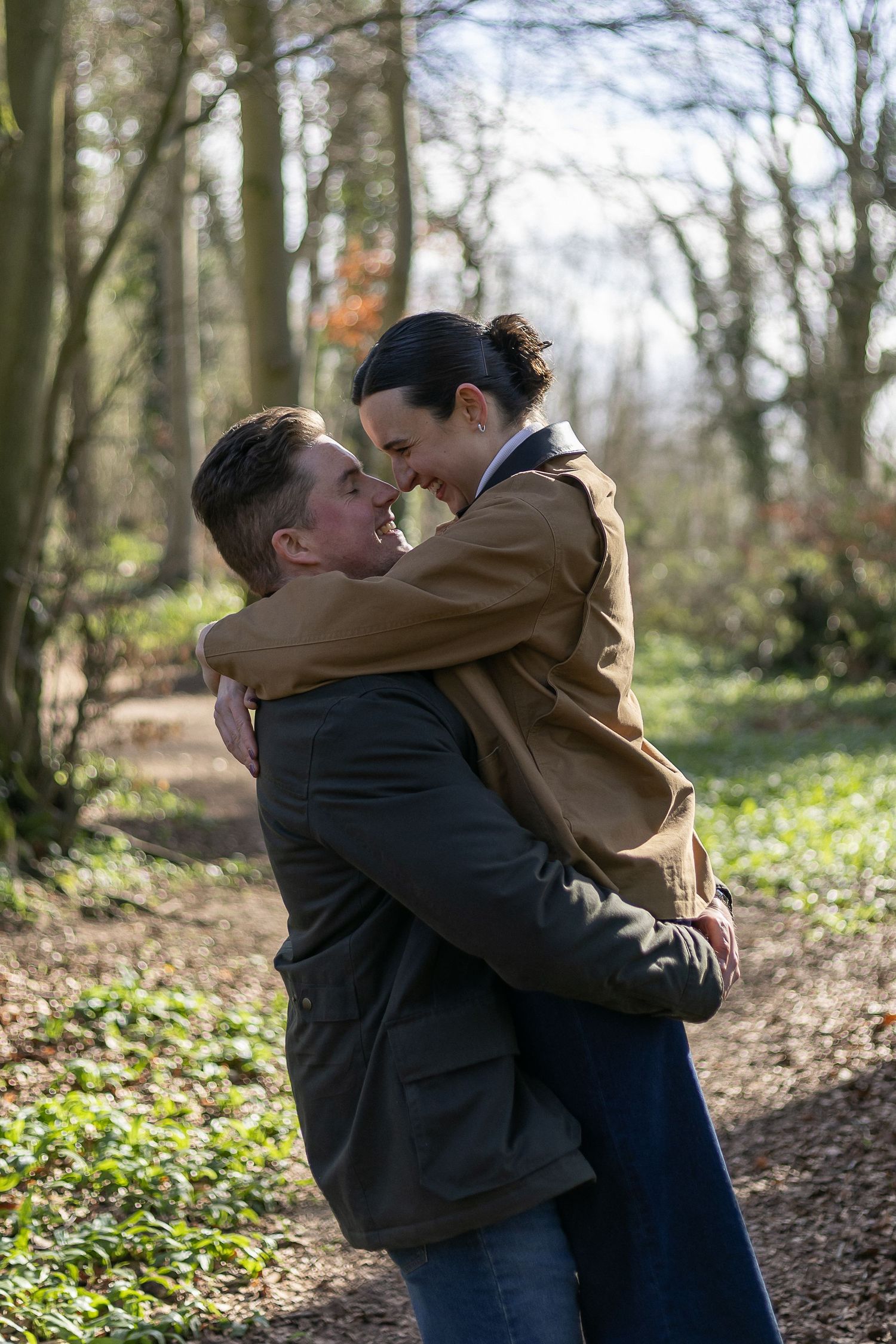 A couple embraces warmly in a sunlit wooded area surrounded by fallen autumn leaves and trees.
