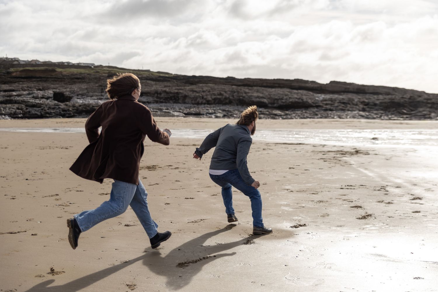 Two people playfully chase each other along a sandy beach on a cloudy day.