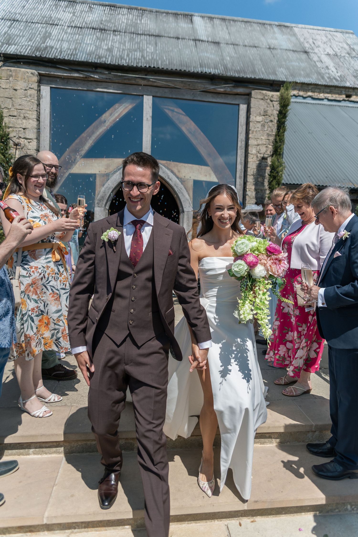 Newlywed couple exits wedding venue holding hands while smiling guests throw confetti on a sunny day.