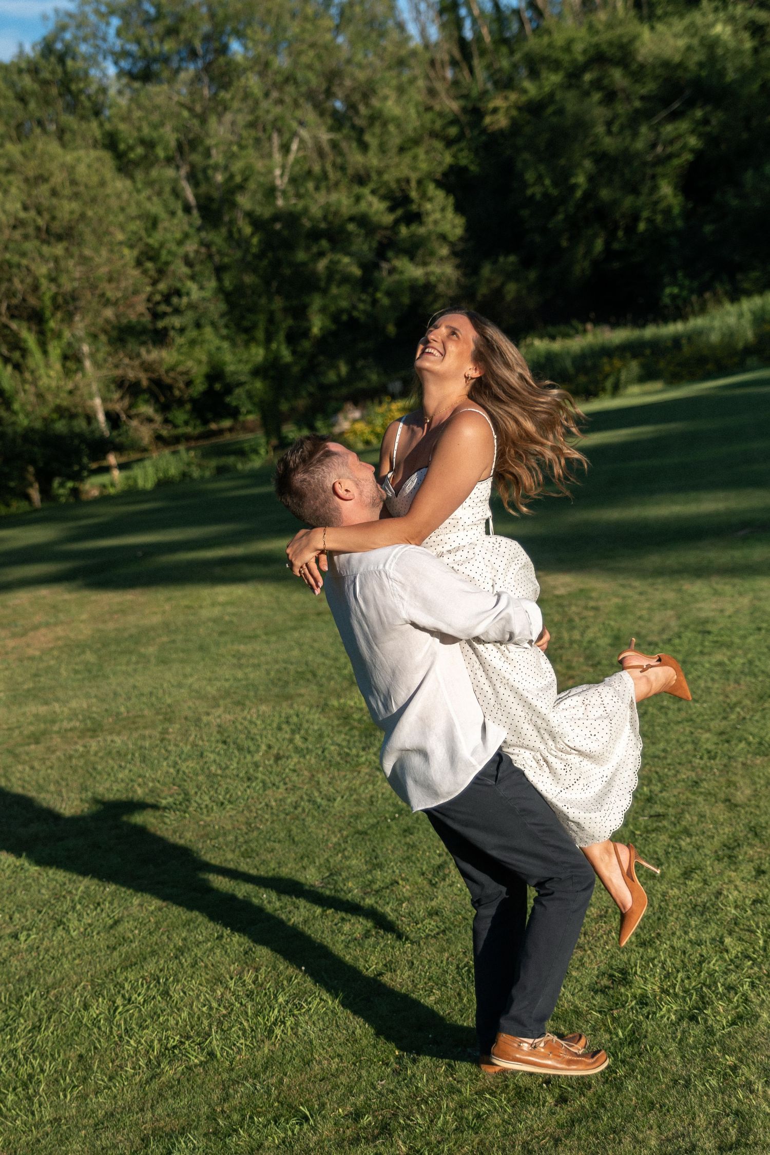 A joyful couple embraces outdoors, man lifting laughing woman in a sunny green park setting.